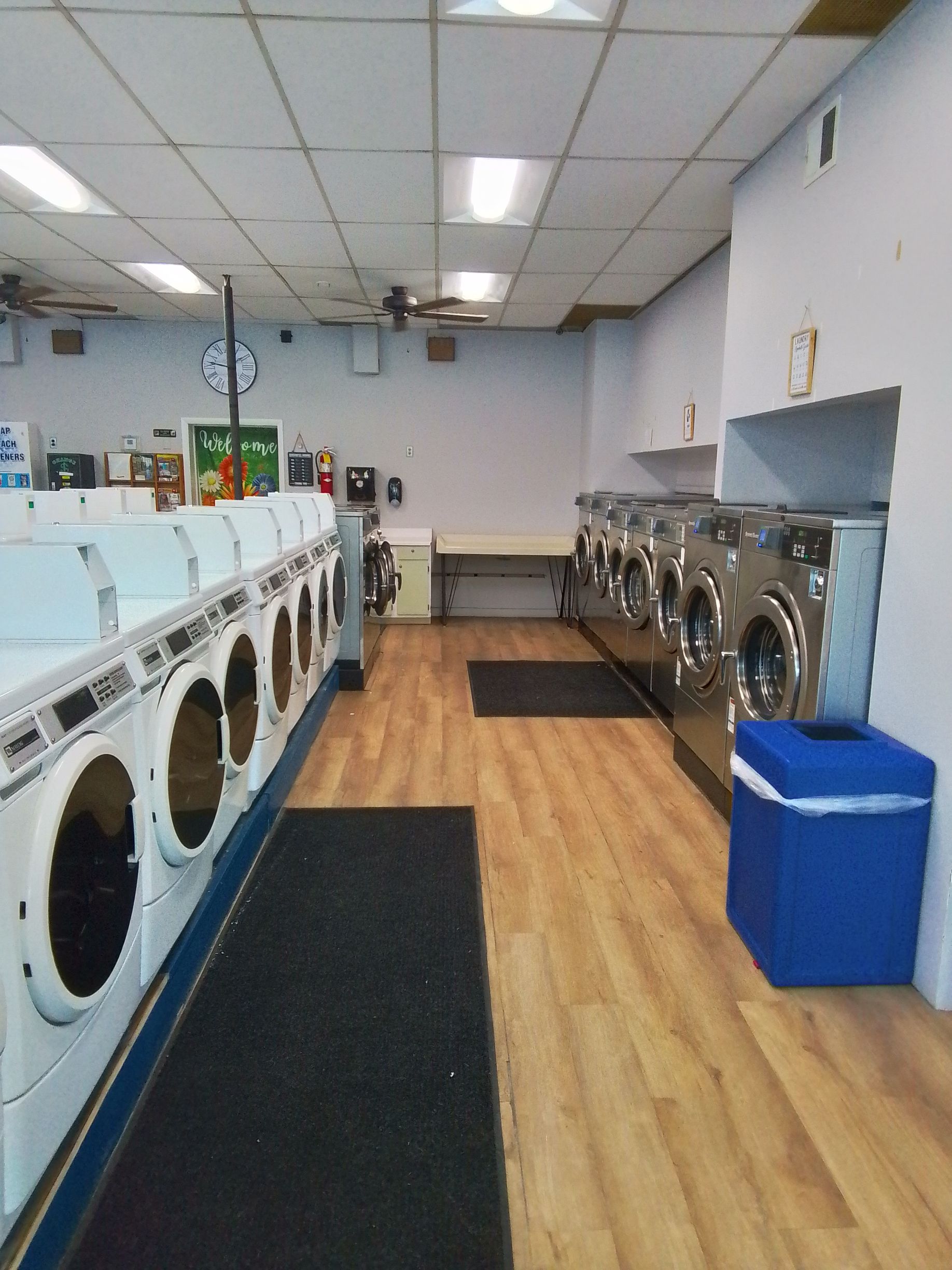 An interior view of a laundromat with rows of washers along the walls, wood-look flooring, and a blue trash bin.