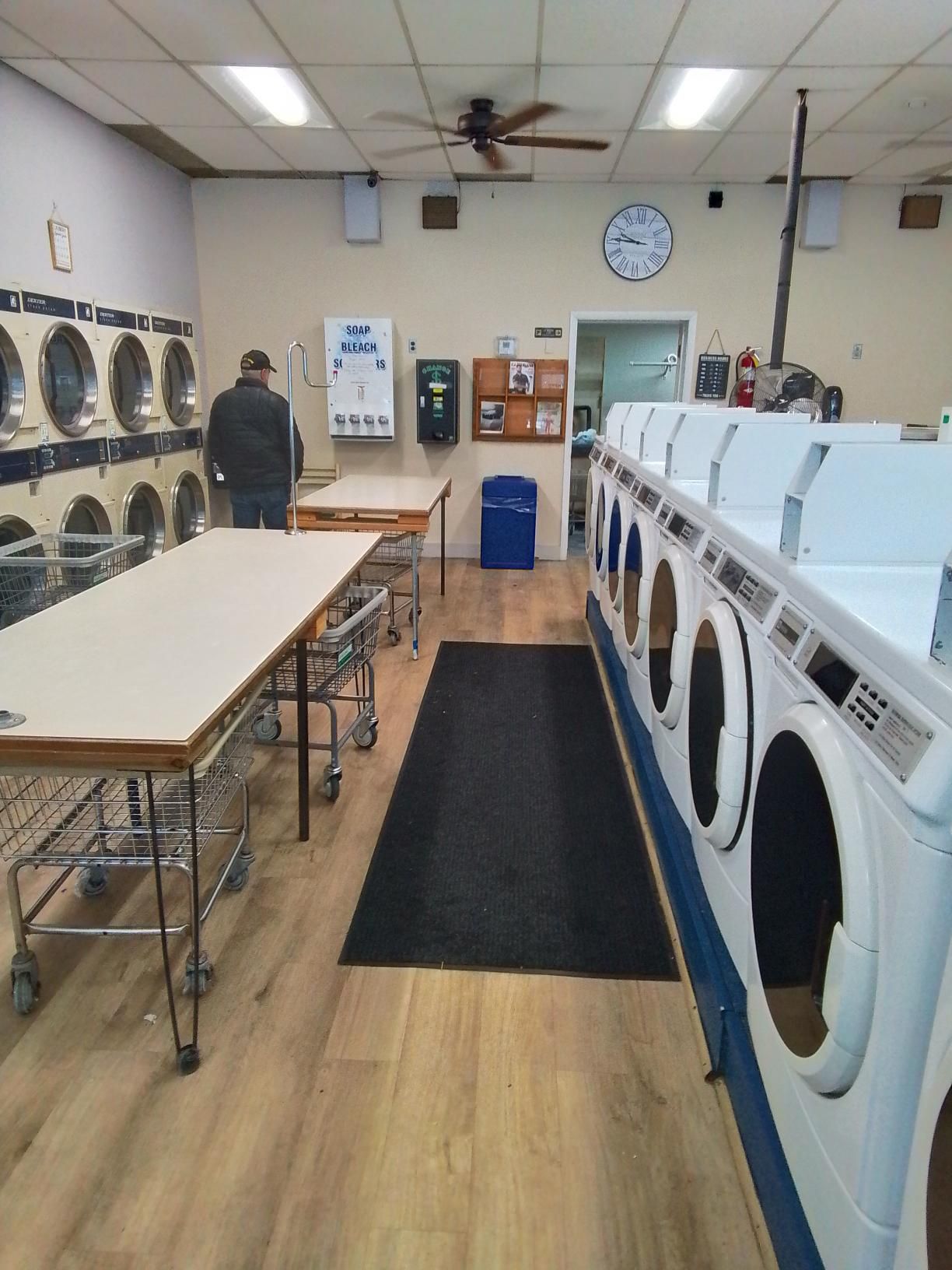A person stands in a laundromat with rows of white dryers, a folding table, and a black floor mat under bright lights.