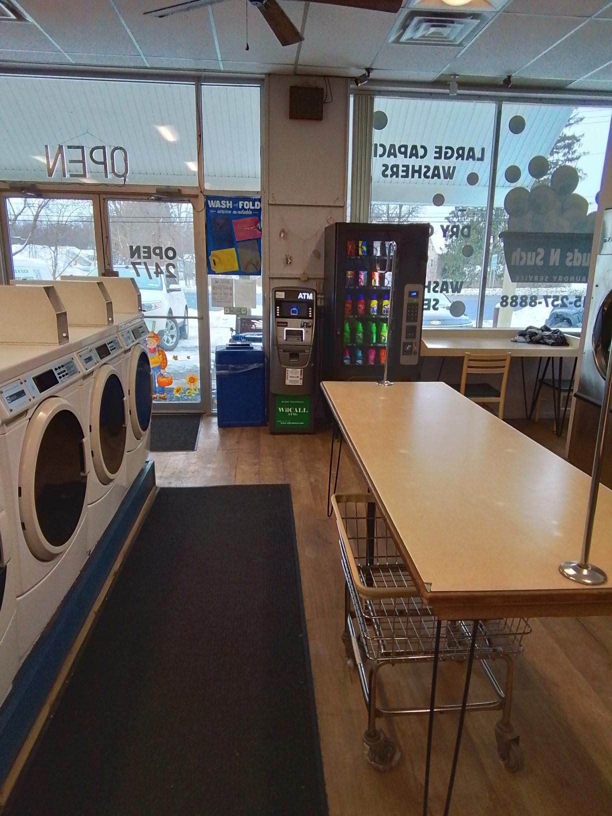 Interior of a laundromat with rows of white washing machines, a long folding table, a vending machine, and a blue bin.