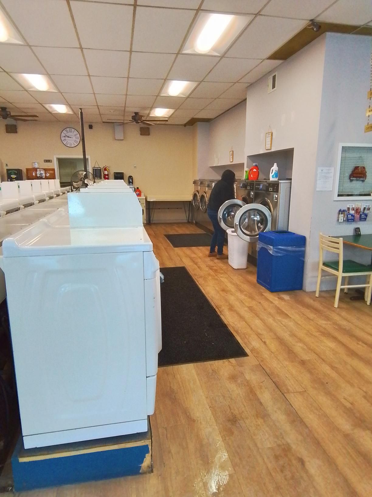 A person loads a washing machine in a laundry room with wood-look flooring, white machines, and a blue bin.
