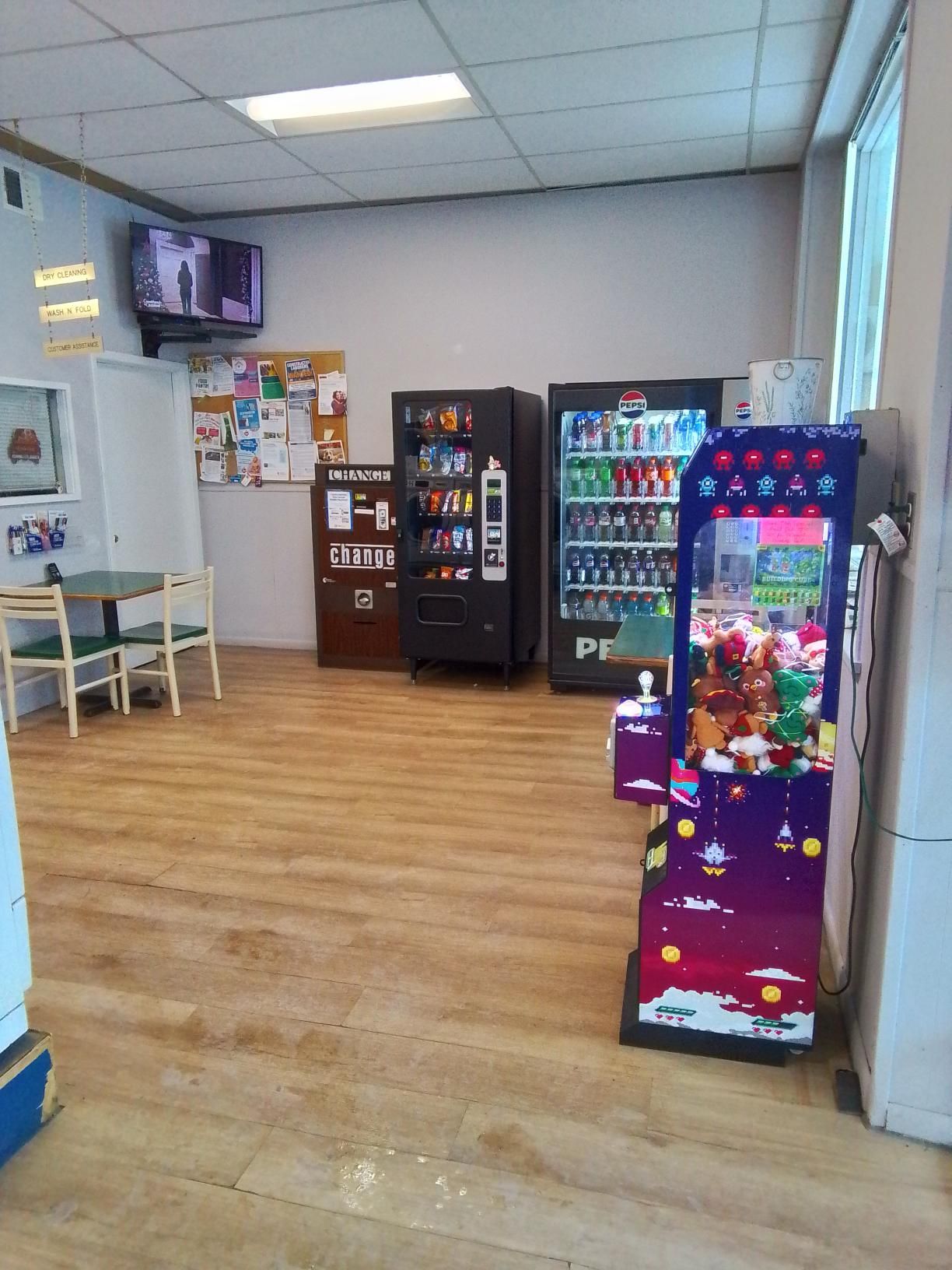 A room with wood-look floors featuring two vending machines, a purple claw machine, and a small table with two chairs.