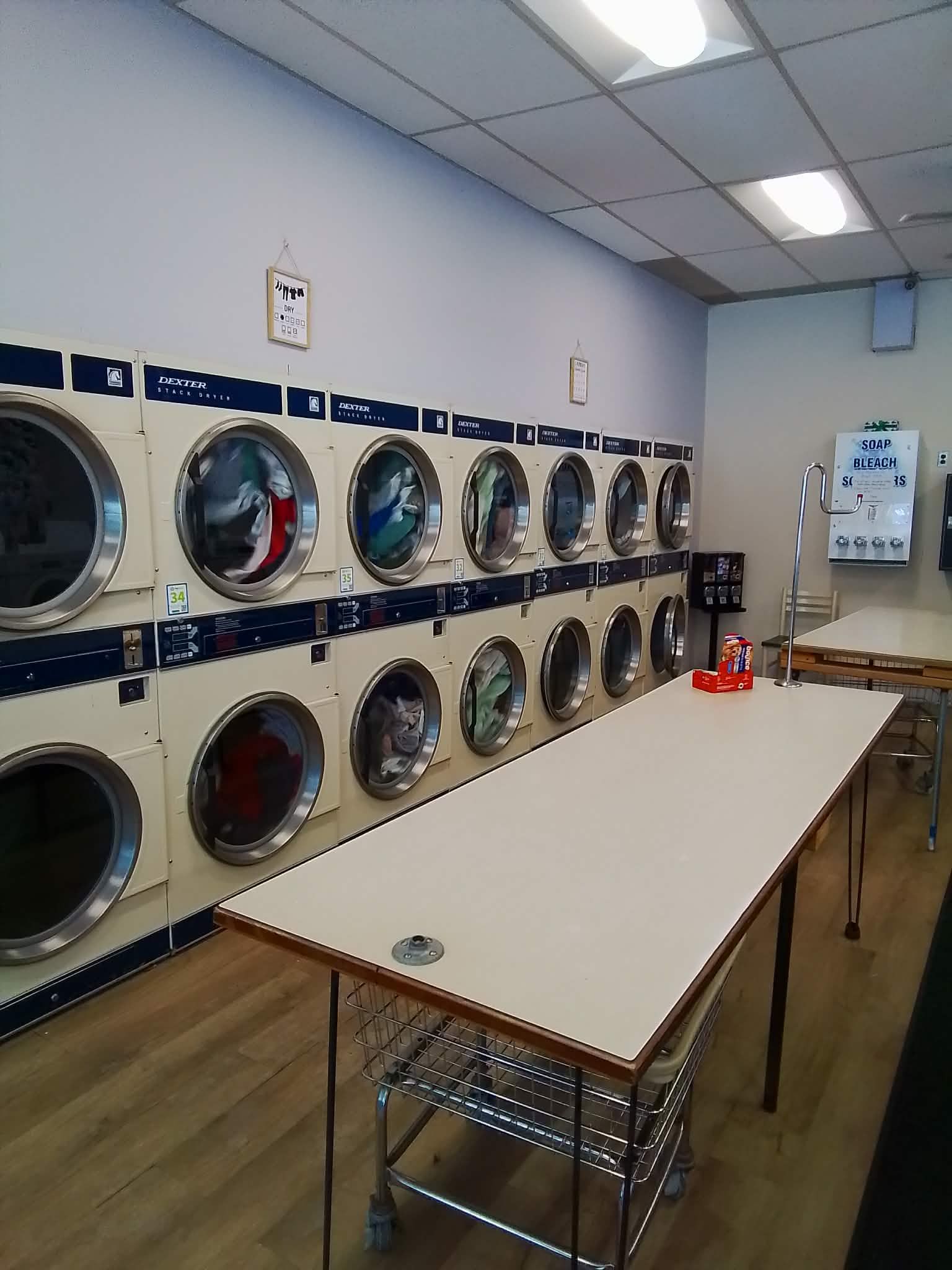 A laundromat with two rows of clothes dryers along the wall and a central folding table on a wood-look floor.