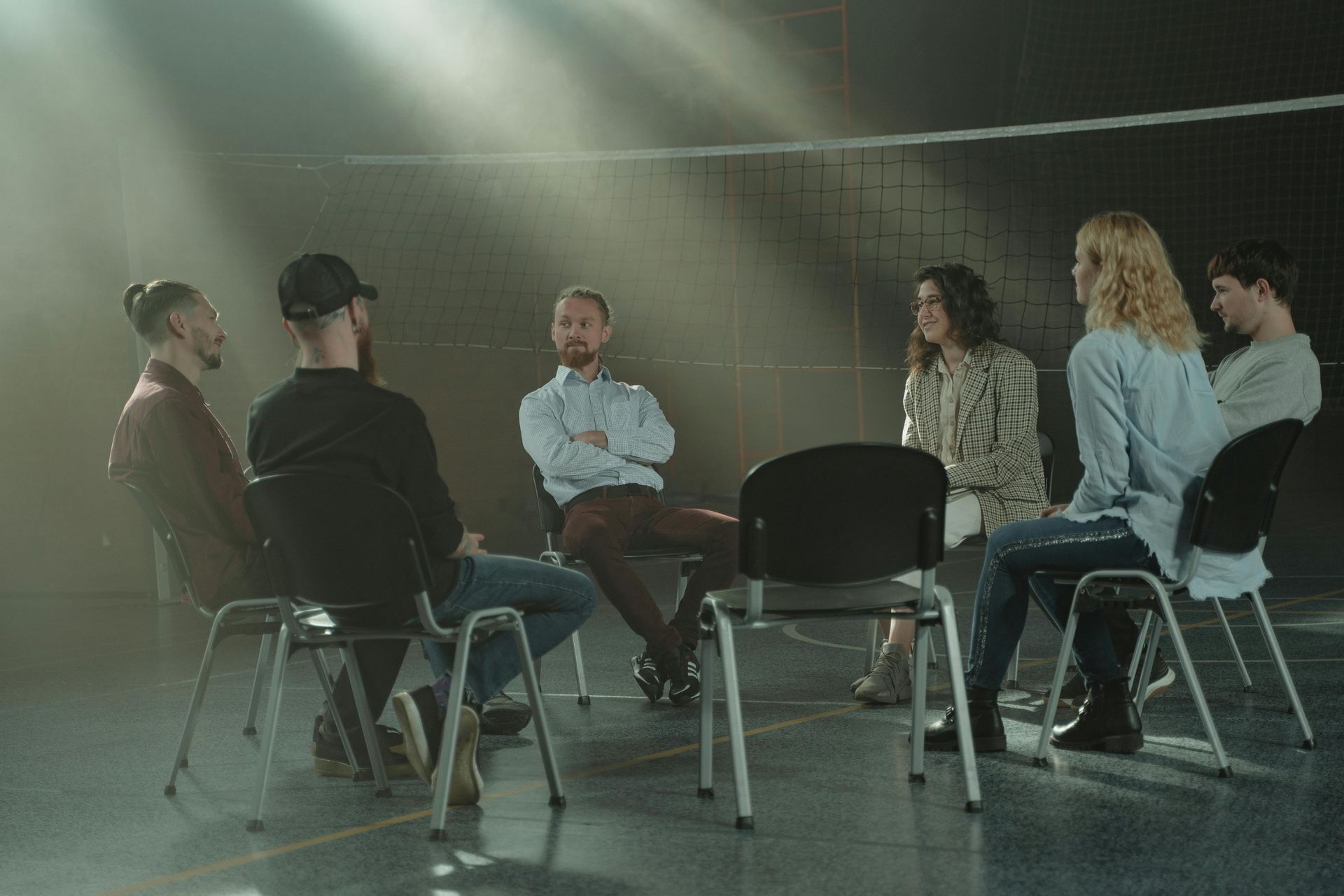 A group of six people sit in a circle, discussing. The setting is indoors, with a net visible.