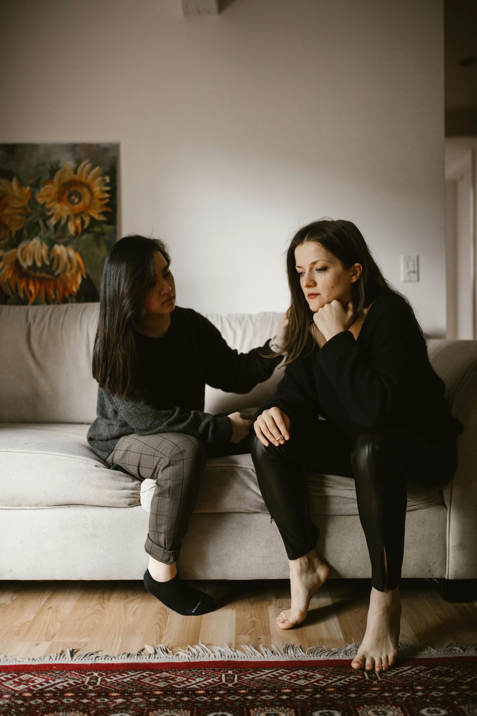 Two women on a couch, one comforts the other, who looks sad. Living room with art and rug.