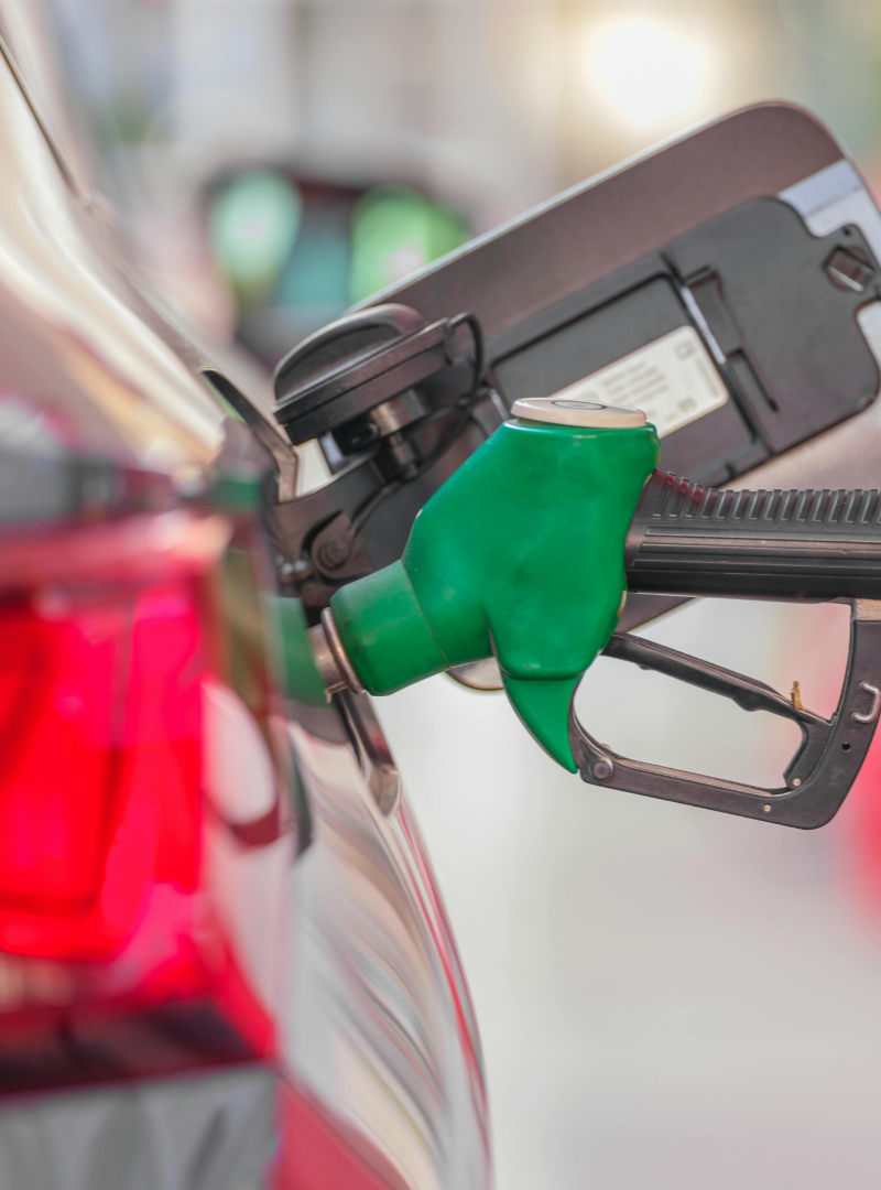 Green fuel nozzle inserted into a car’s gas tank at a gas station, with blurred red tail lights in the background