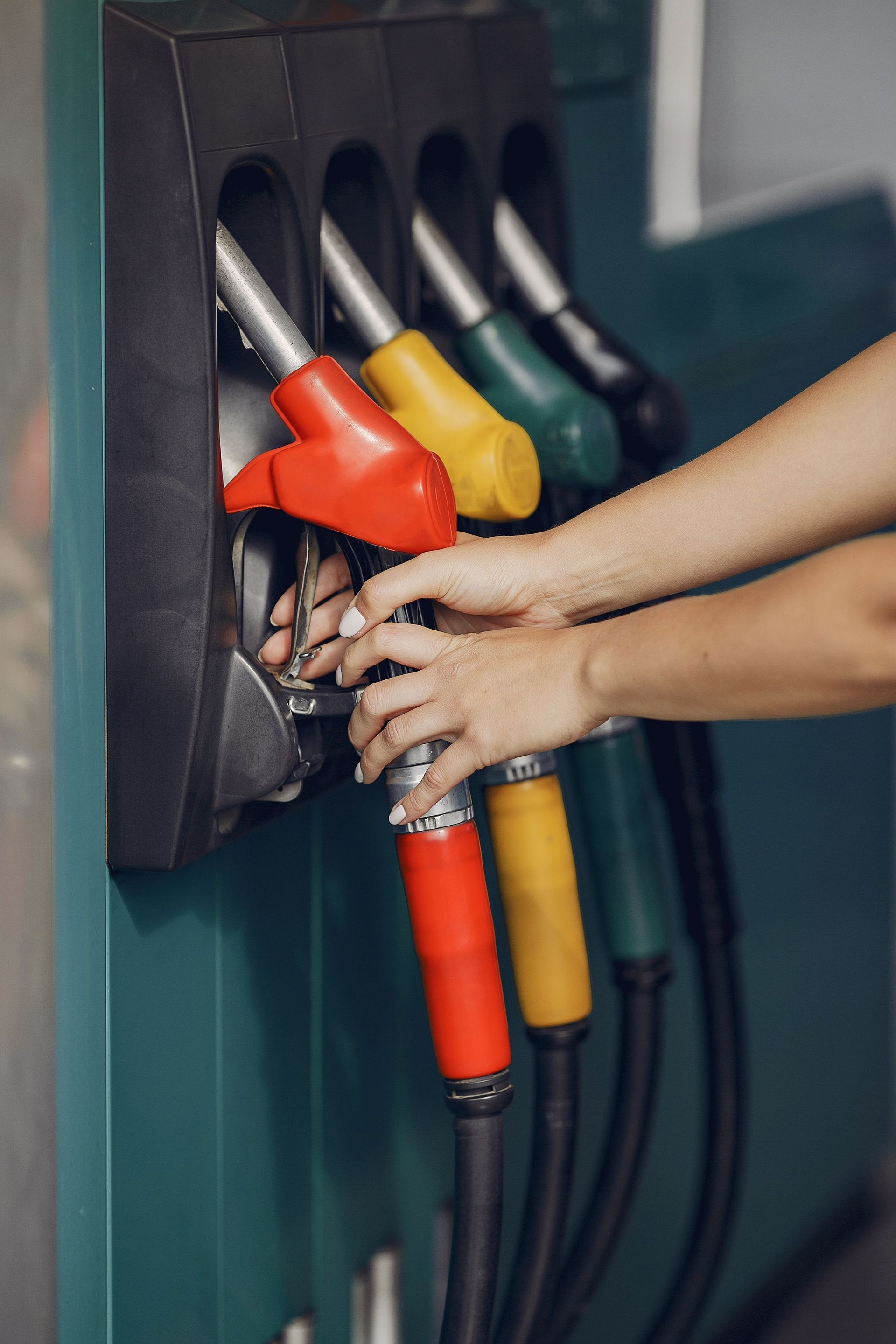 Hands refueling a car at a gas station, holding red, yellow, and green fuel nozzles.