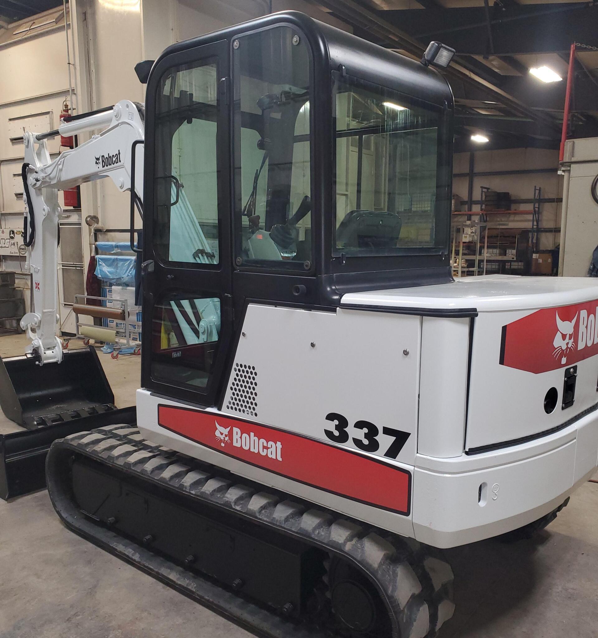 White Bobcat 337 mini excavator on tracks, indoors. Black cab, arm extended with a bucket.
