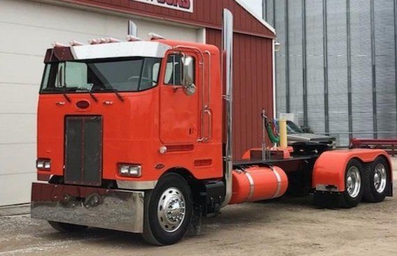 Orange Peterbilt semi-truck parked in front of a red building, with a silver bumper and fuel tanks.