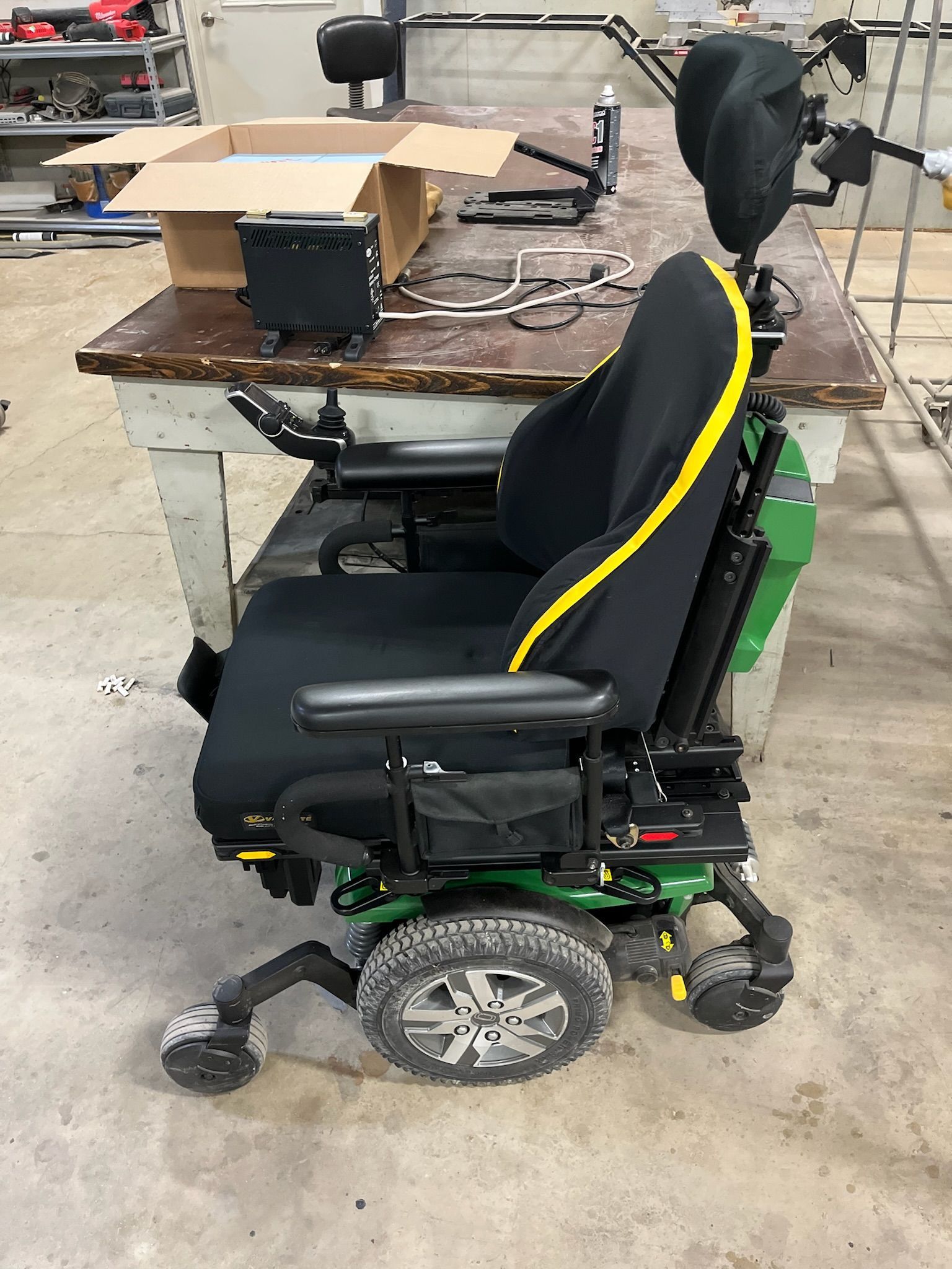 Green and black power wheelchair with black seat cover, parked next to a cluttered workbench.