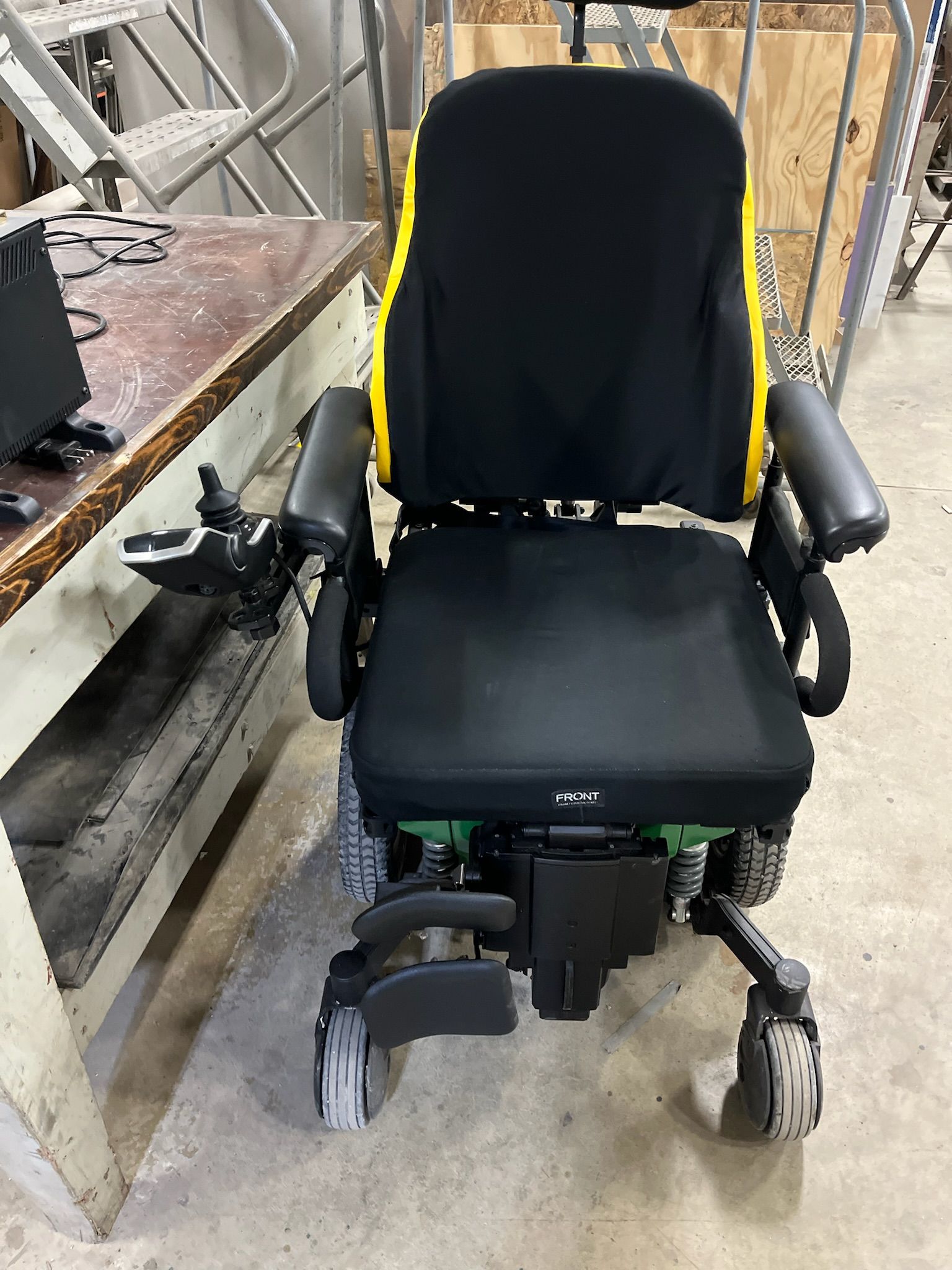 Black and yellow electric wheelchair on concrete floor with a control panel and armrests.