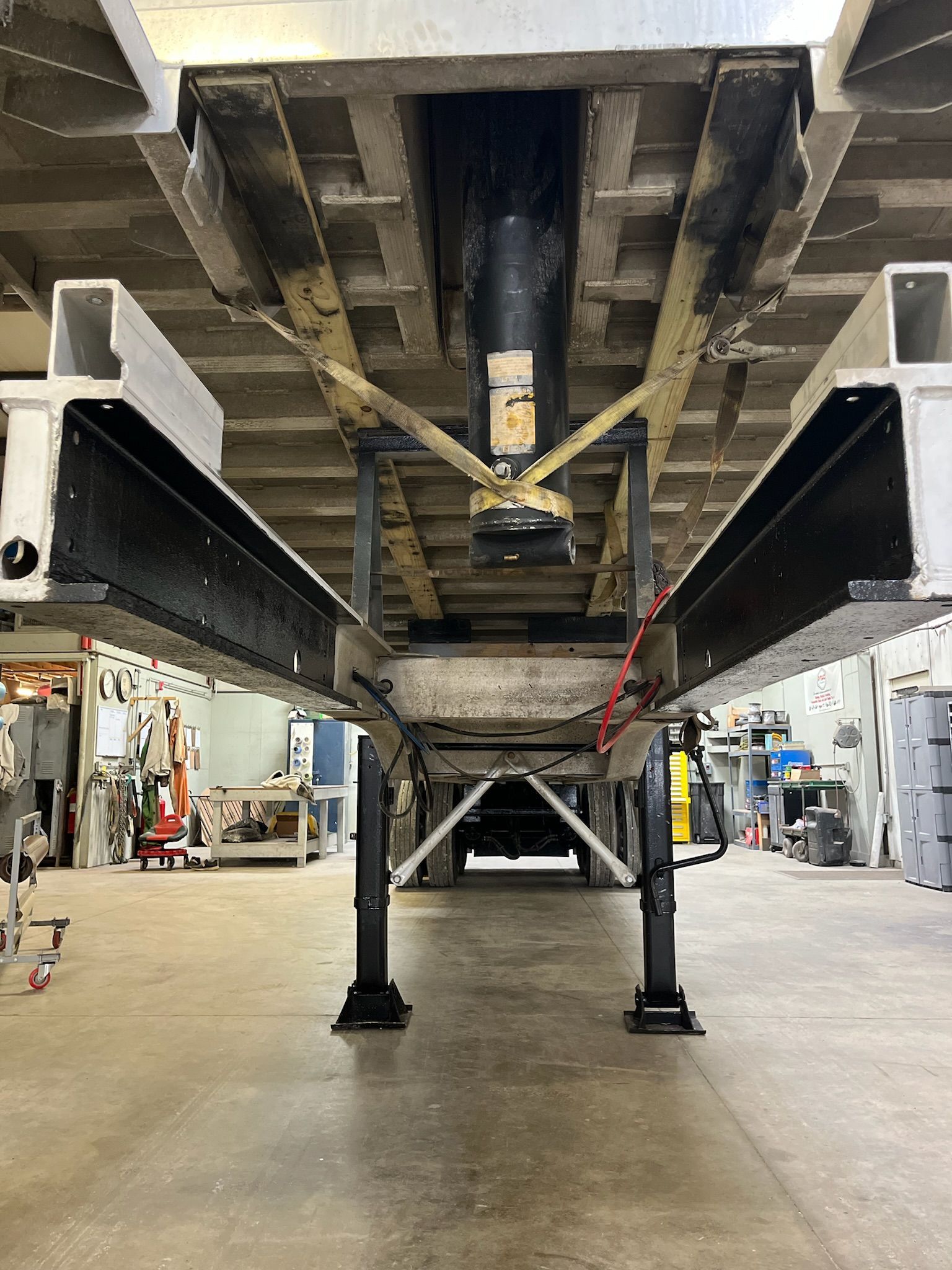 Underside view of a trailer chassis supported by black jacks. Steel beams and crossbars are visible in a garage setting.