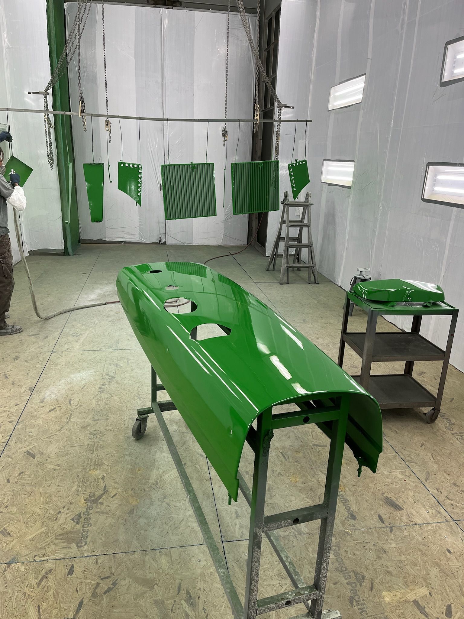 A green painted tractor hood in a paint booth. A person sprays paint; other green parts hang.