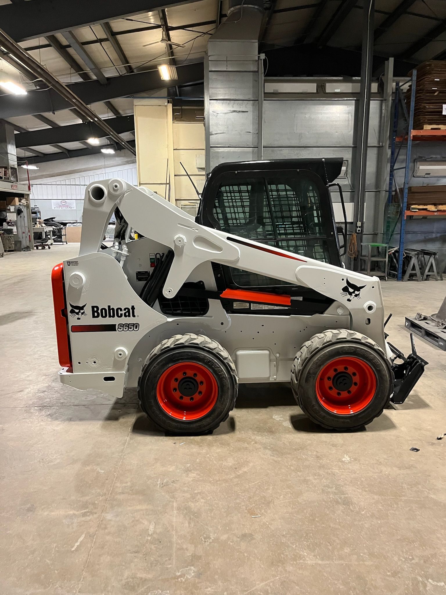 White and orange Bobcat skid-steer loader parked indoors with orange wheels.