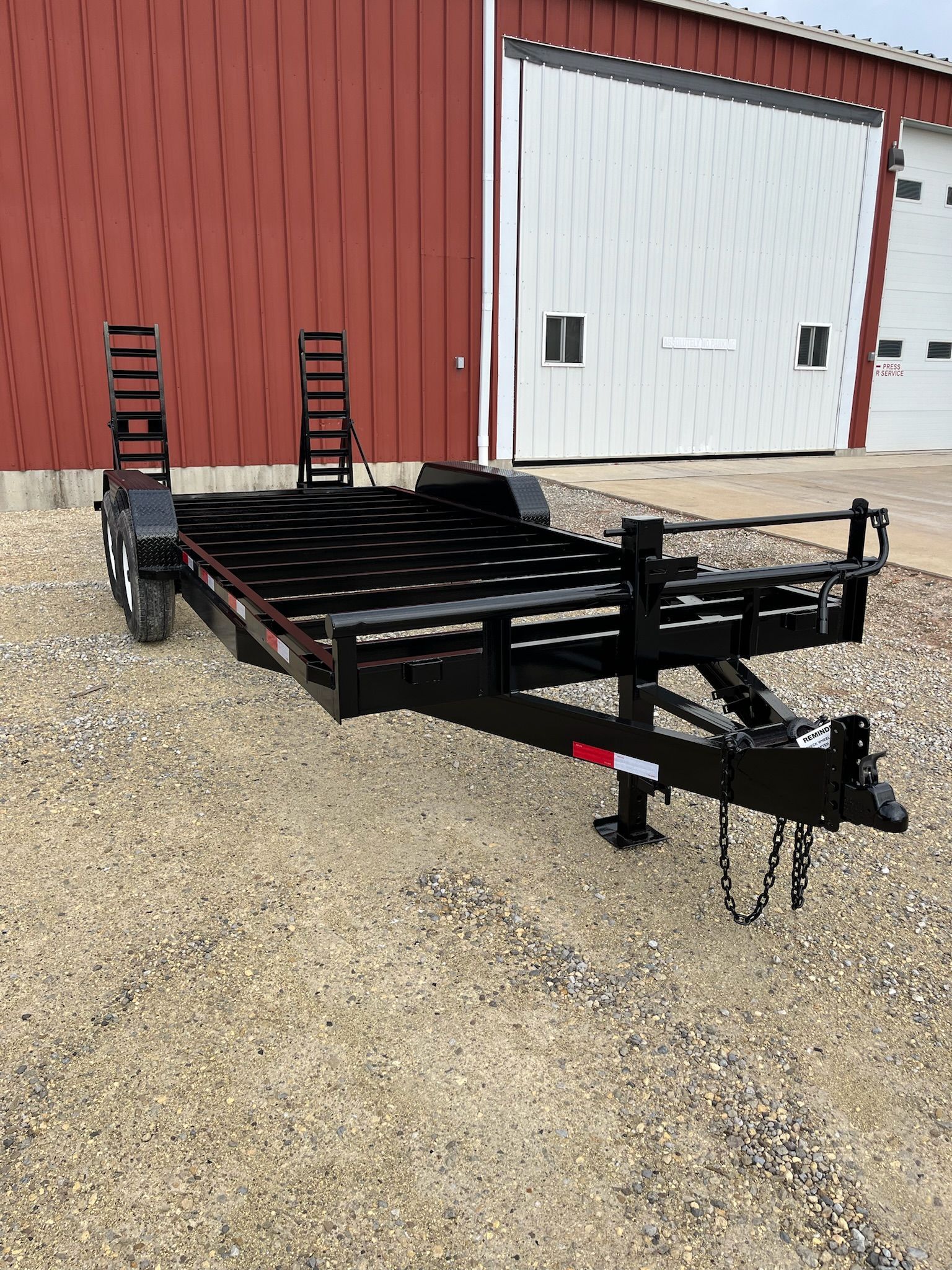 Black utility trailer parked on gravel in front of a red building and a white garage door.