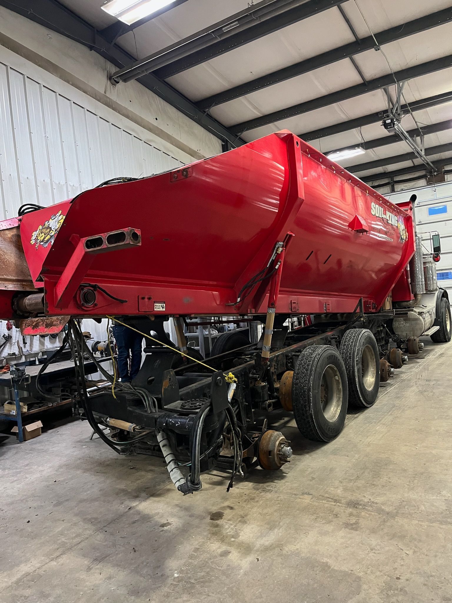 Red dump truck trailer inside a garage, with a raised bed and multiple axles.