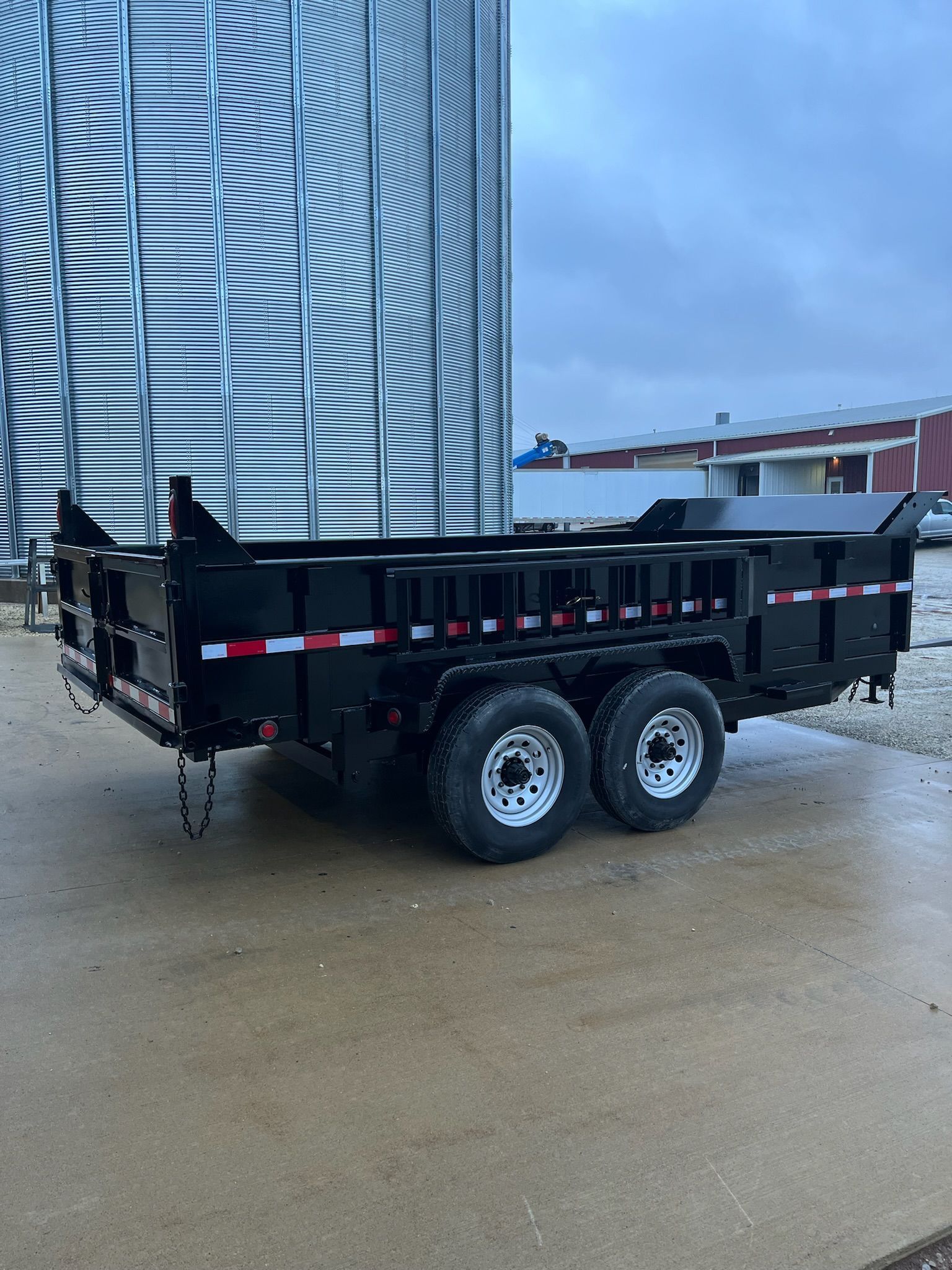 Black dump trailer with dual wheels, parked on wet concrete, near a metal grain silo.