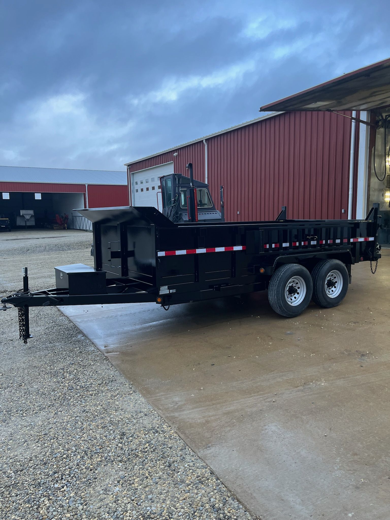 Black dump trailer with dual wheels, parked on gravel in front of a red building and cloudy sky.