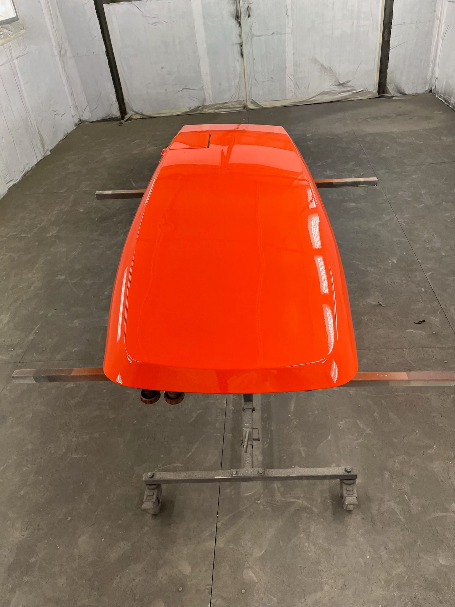 Orange vehicle hood, freshly painted, on a wheeled stand in a workshop.