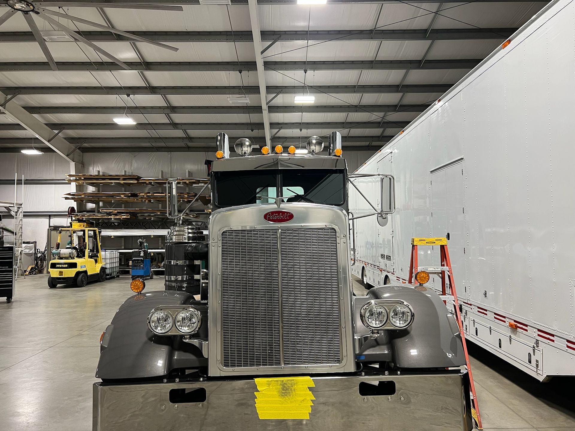 A gray semi-truck parked inside a warehouse, with a forklift and trailer visible in the background.