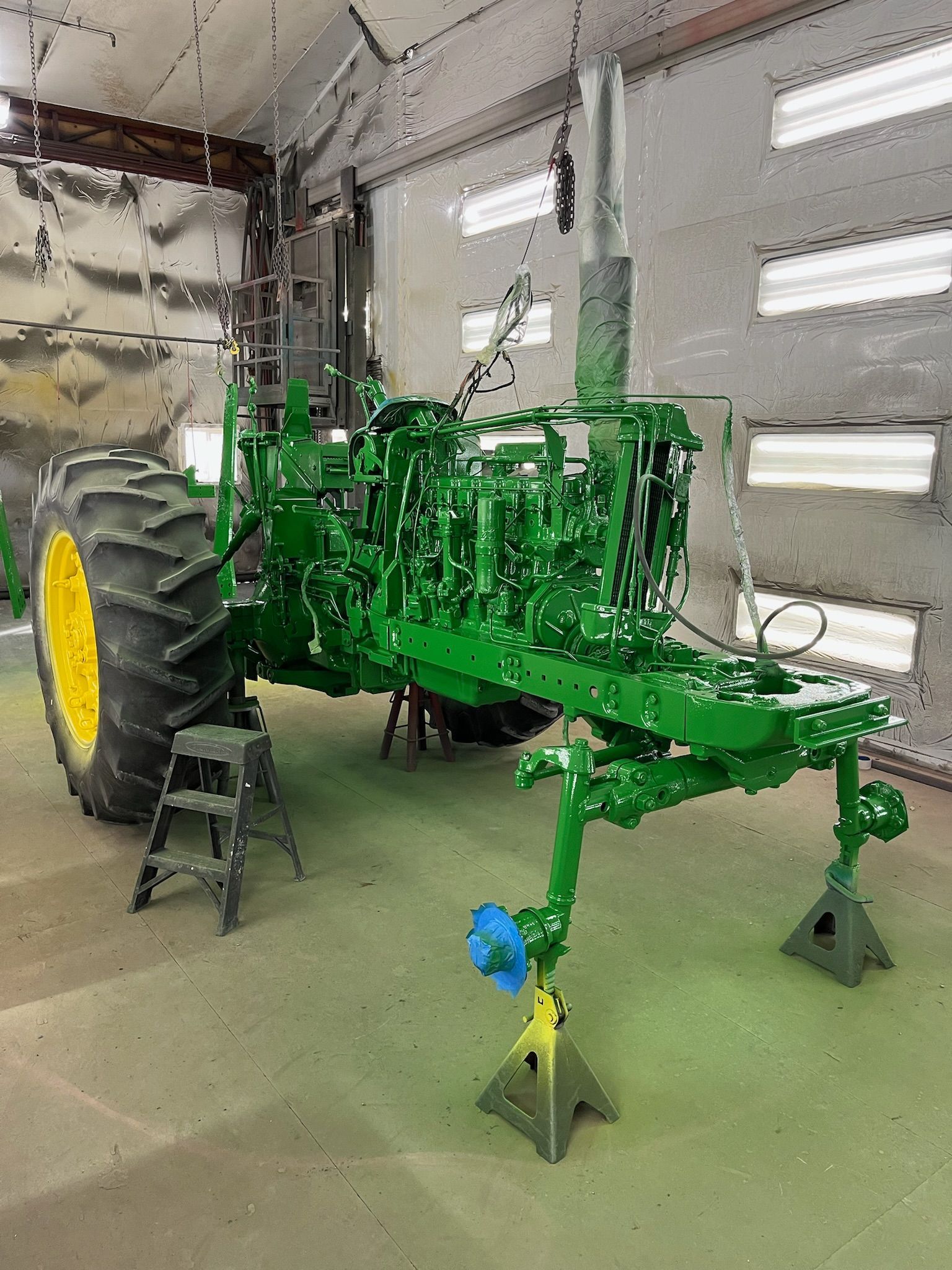 Green John Deere tractor on jack stands in a repair shop, recently painted.