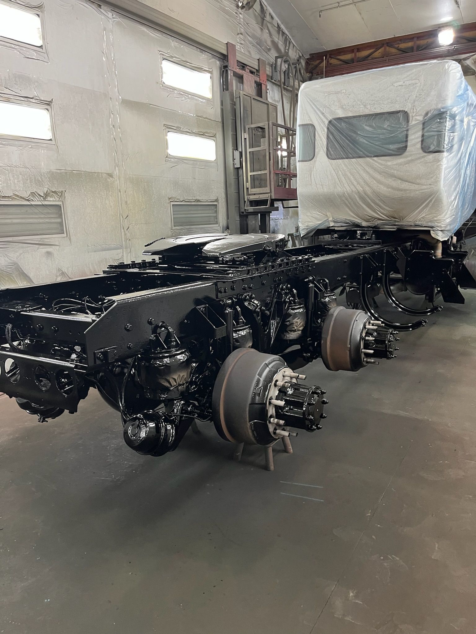 Black truck chassis in a workshop, white truck cab wrapped in plastic.