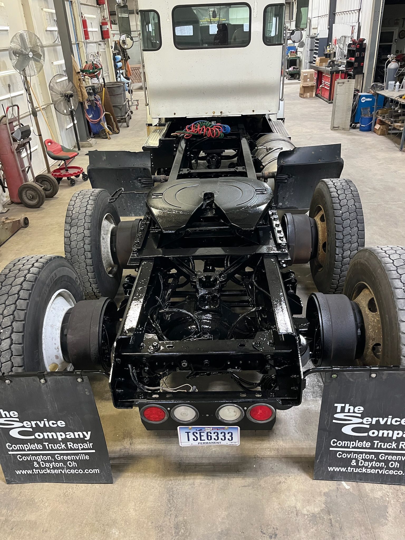 Undercarriage of a truck cab, freshly painted black. Tires, lights, and trailer hitch visible inside a shop.