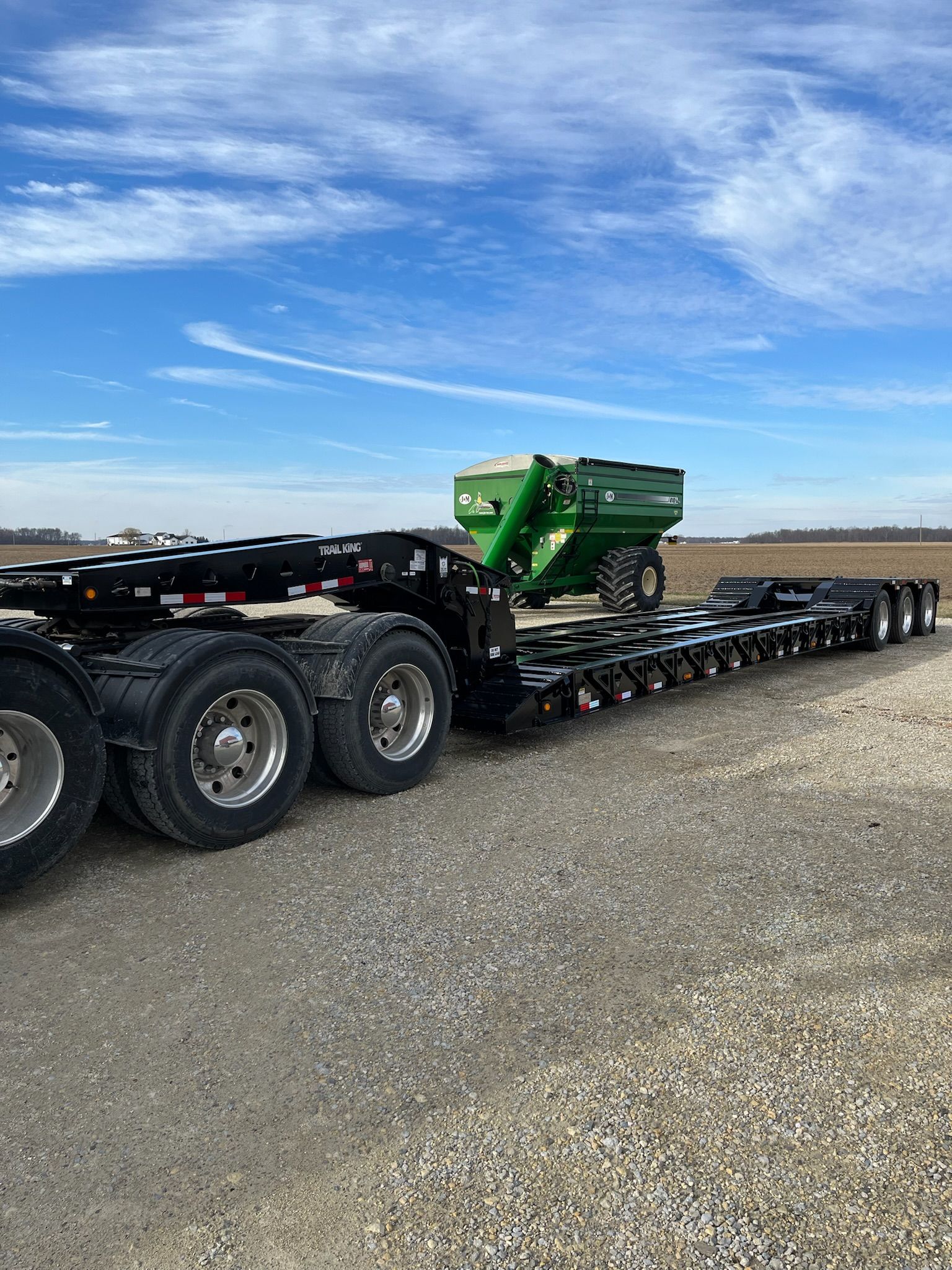 Black flatbed trailer hauling a green grain cart on a gravel road under a blue sky.