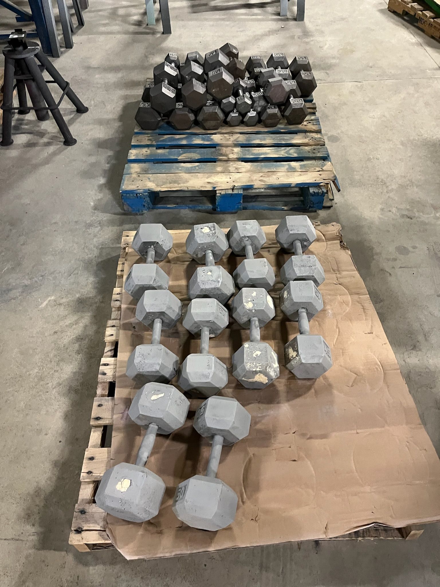 Two pallets of dumbbells in a gym setting; one pallet has dark weights, the other has gray weights.