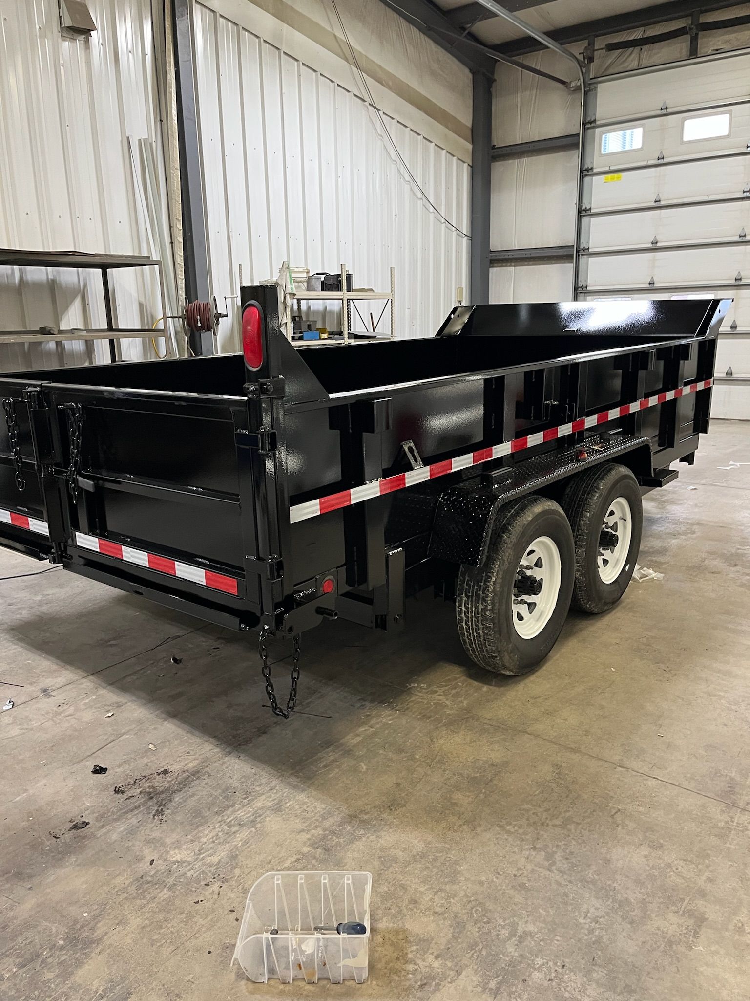 Black dump trailer with white tires, red and white reflective tape, in a workshop.