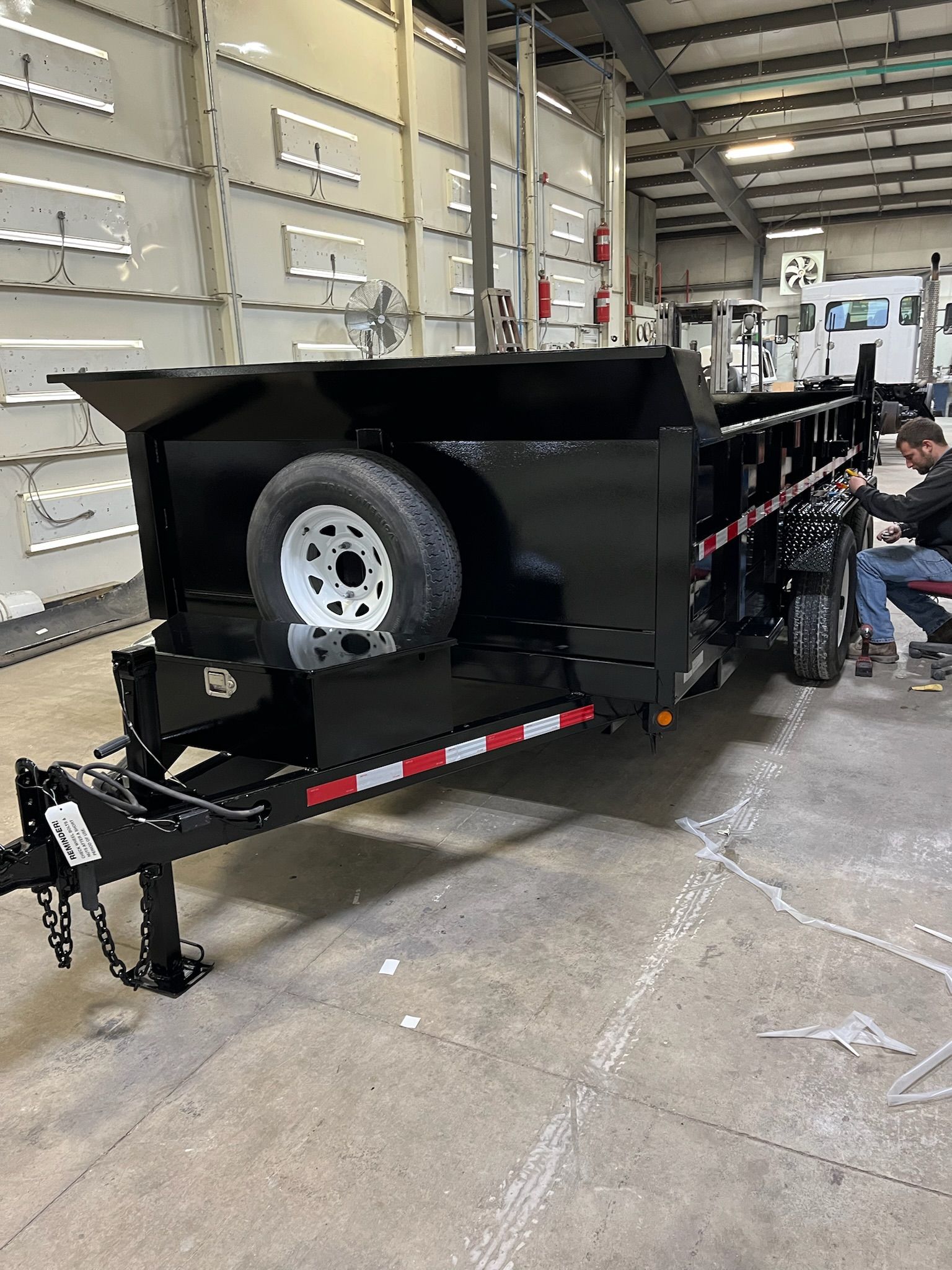 Black dump trailer with a spare tire, parked in a building with a person nearby.