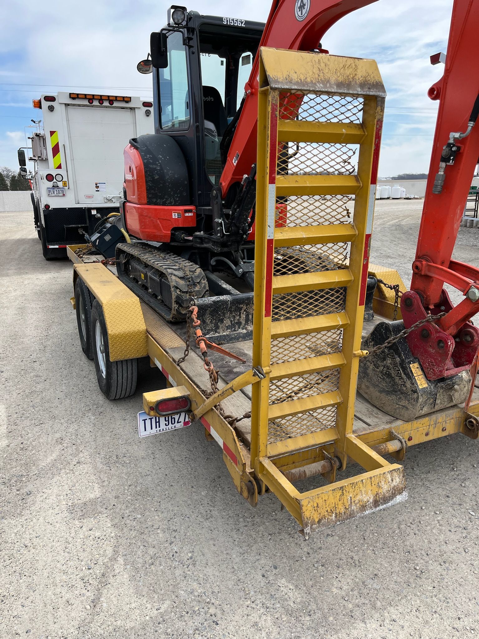 Yellow trailer with mini-excavator and ramp. White truck in the background. Outdoors.