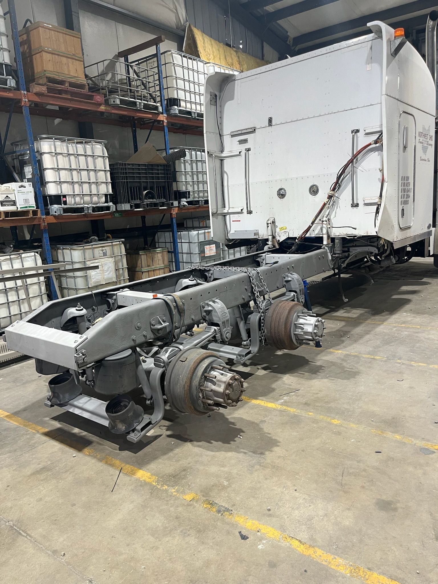 White truck chassis in a warehouse setting, with a cab and wheels, some parts on shelves in the background.