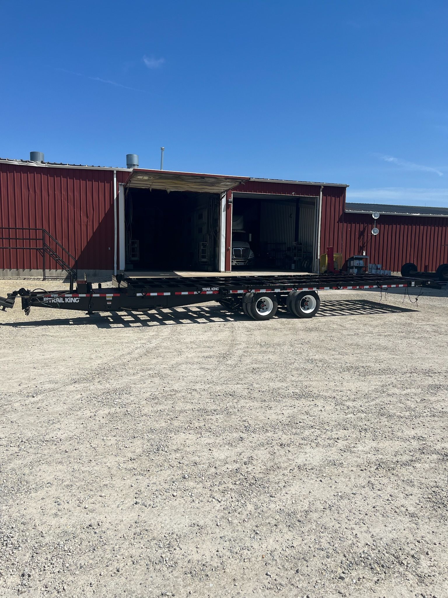 Black trailer parked in front of a red building with open doors, on a gravel lot under a blue sky.