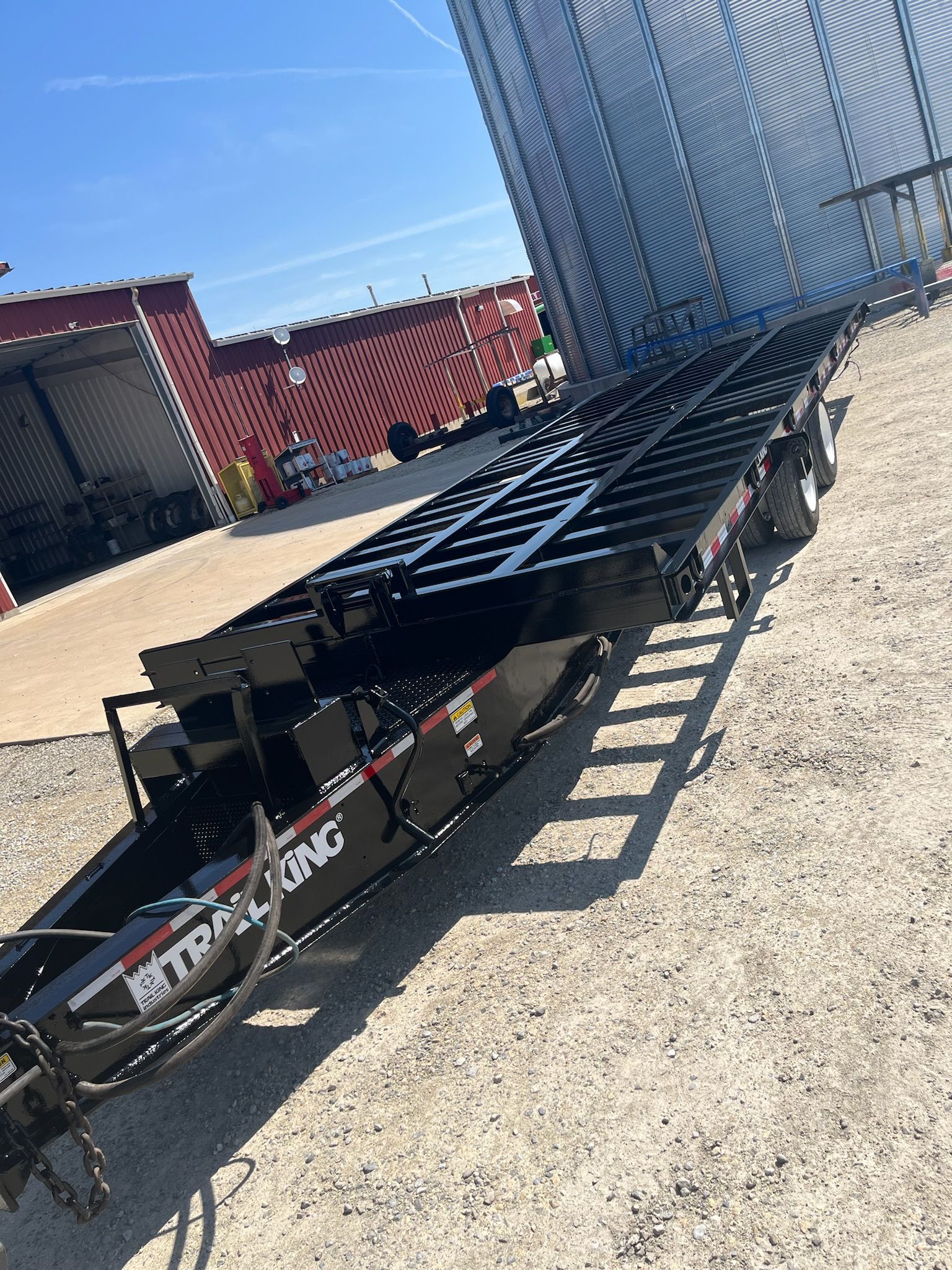 Black flatbed trailer with ramps parked in a gravel lot near a grain silo.