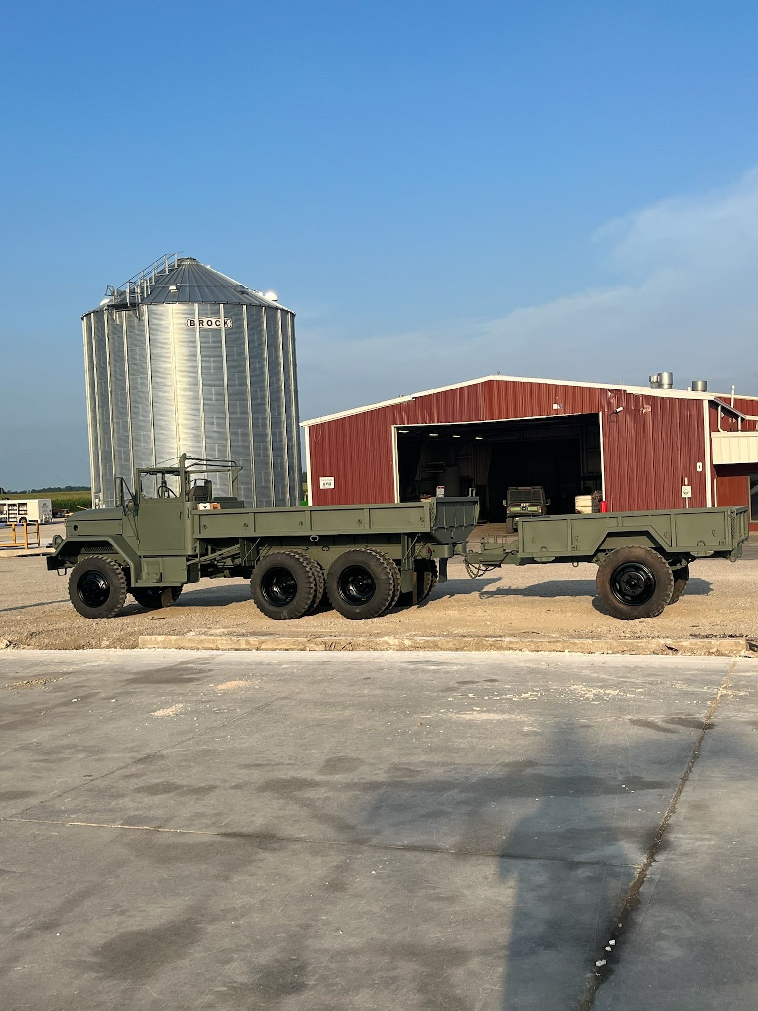 Olive green military truck with trailer on concrete near a grain silo and red barn.