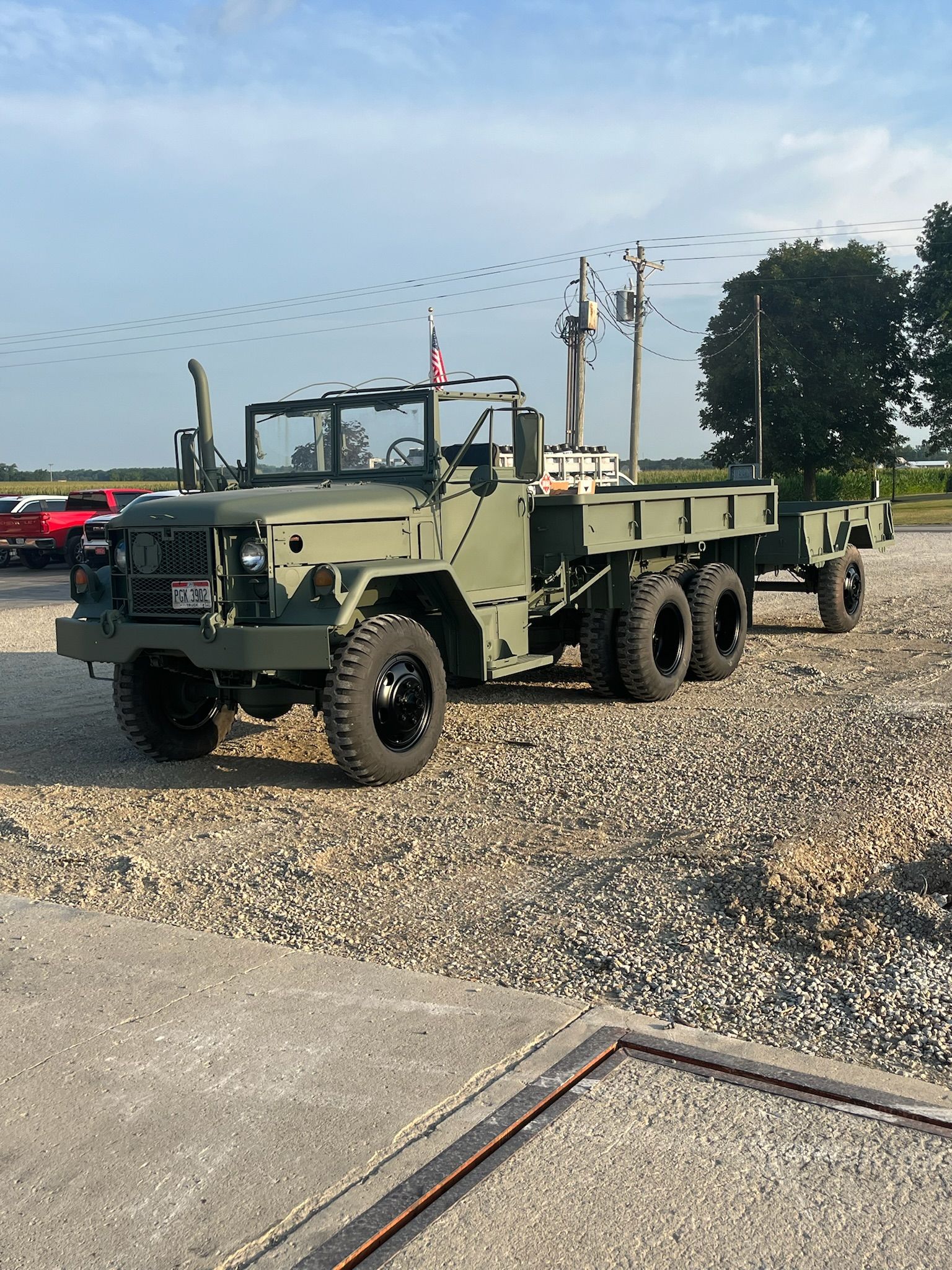Olive green military truck with trailer parked on gravel.