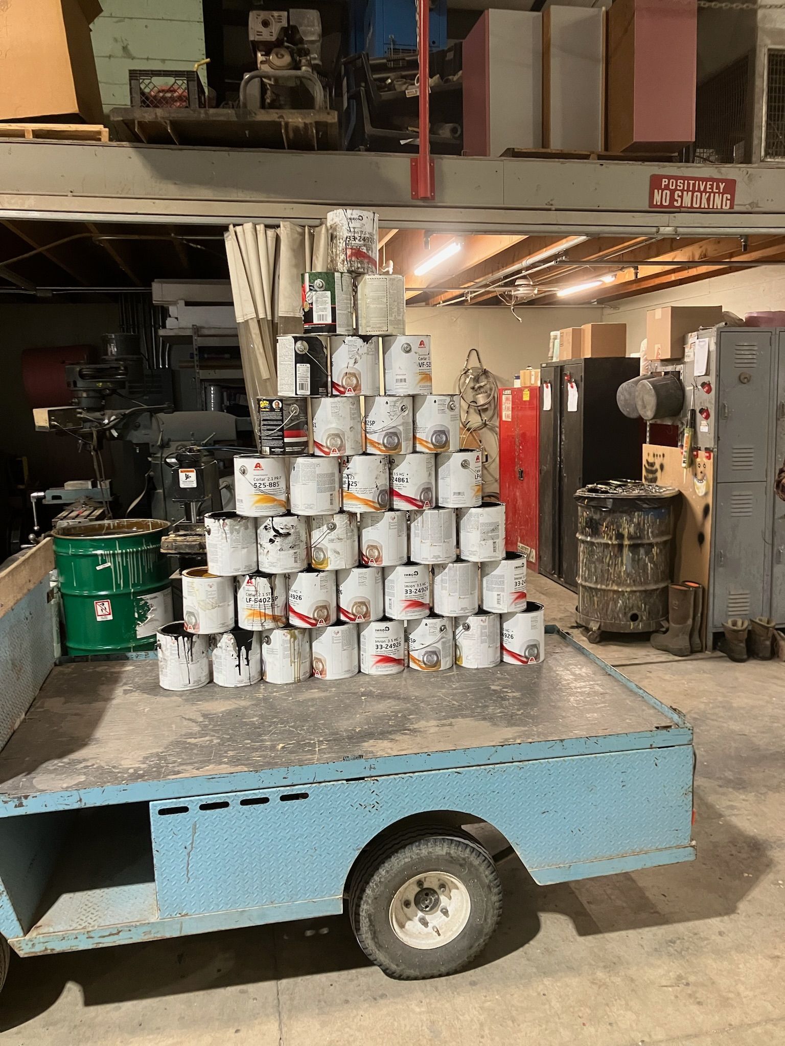 Stack of paint cans on a light blue flatbed trailer inside a warehouse, other equipment visible.