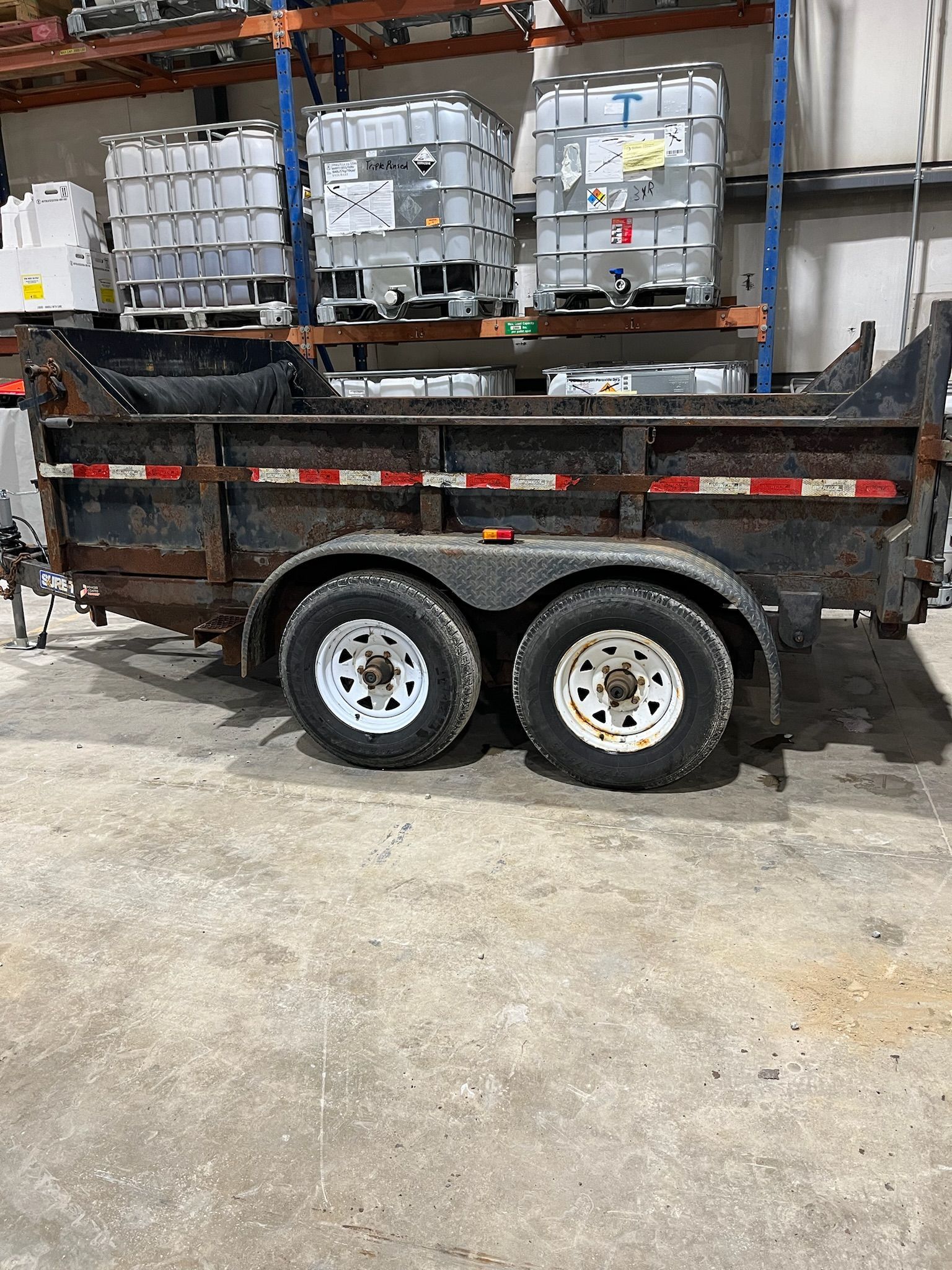 Black dump trailer with white wheels, parked in warehouse, with barrels on shelves in the background.