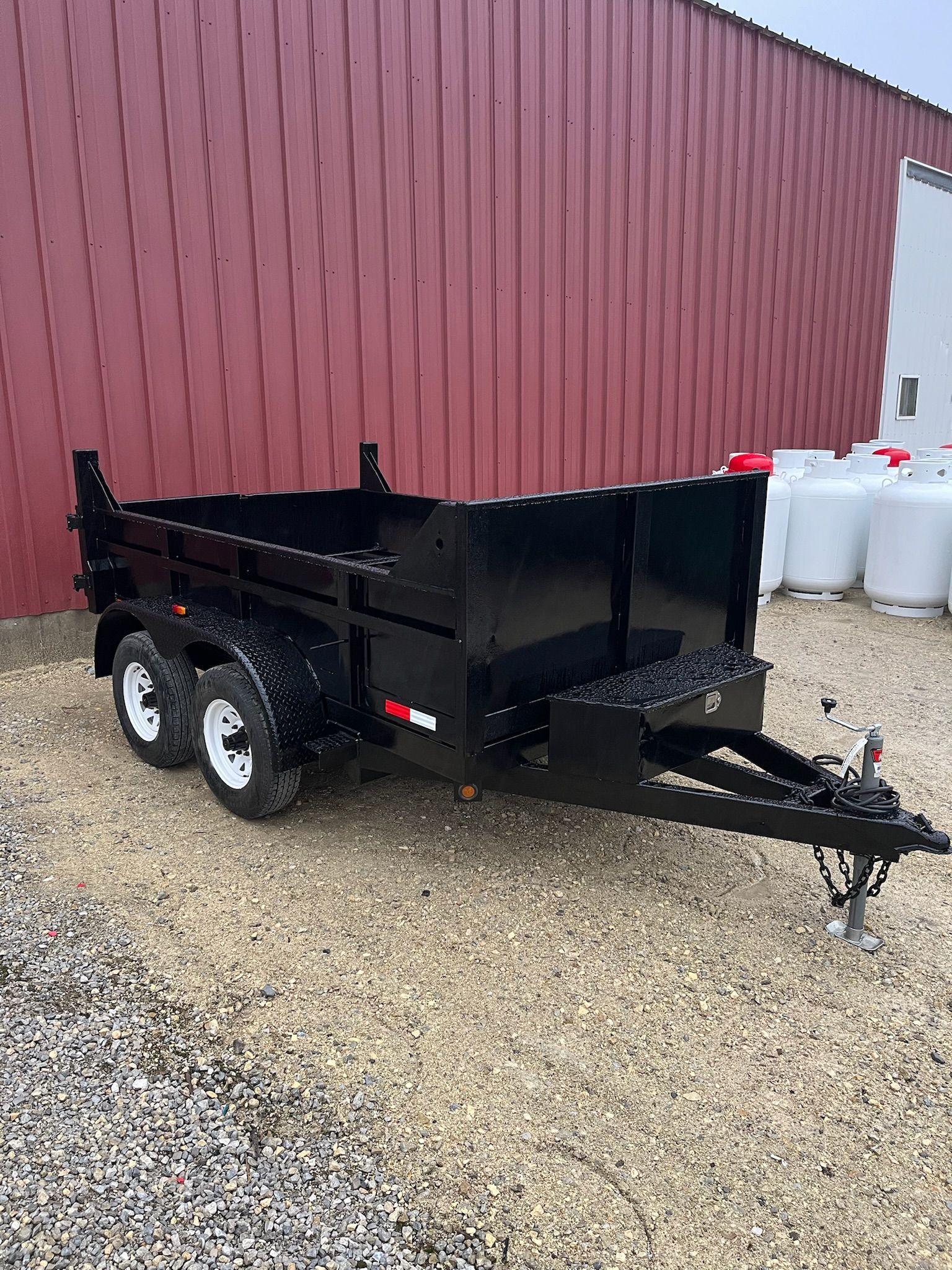 Black dump trailer with white wheels, parked on gravel in front of a red building and propane tanks.