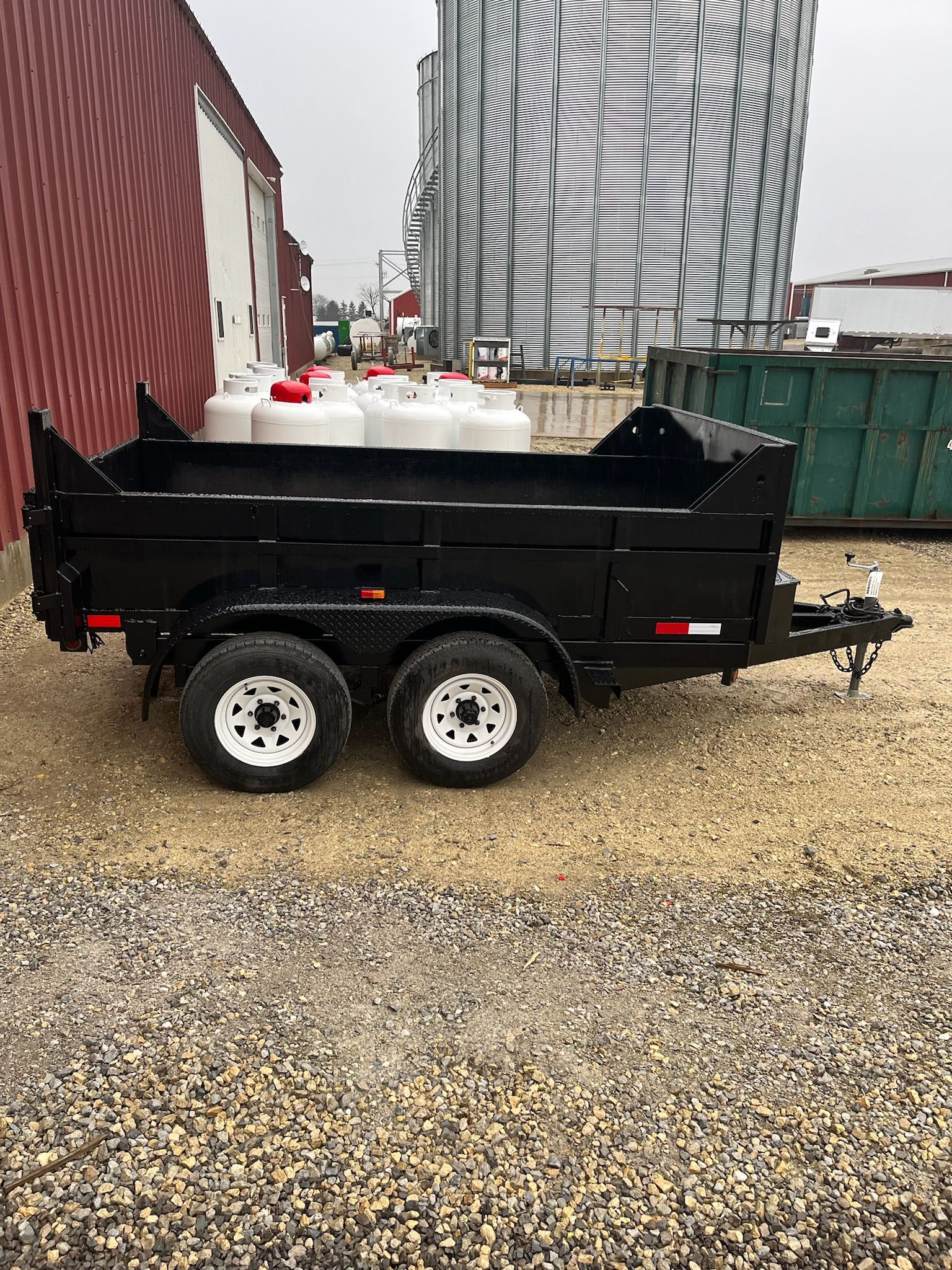 Black dump trailer with white-rimmed tires on gravel, red barn in background.