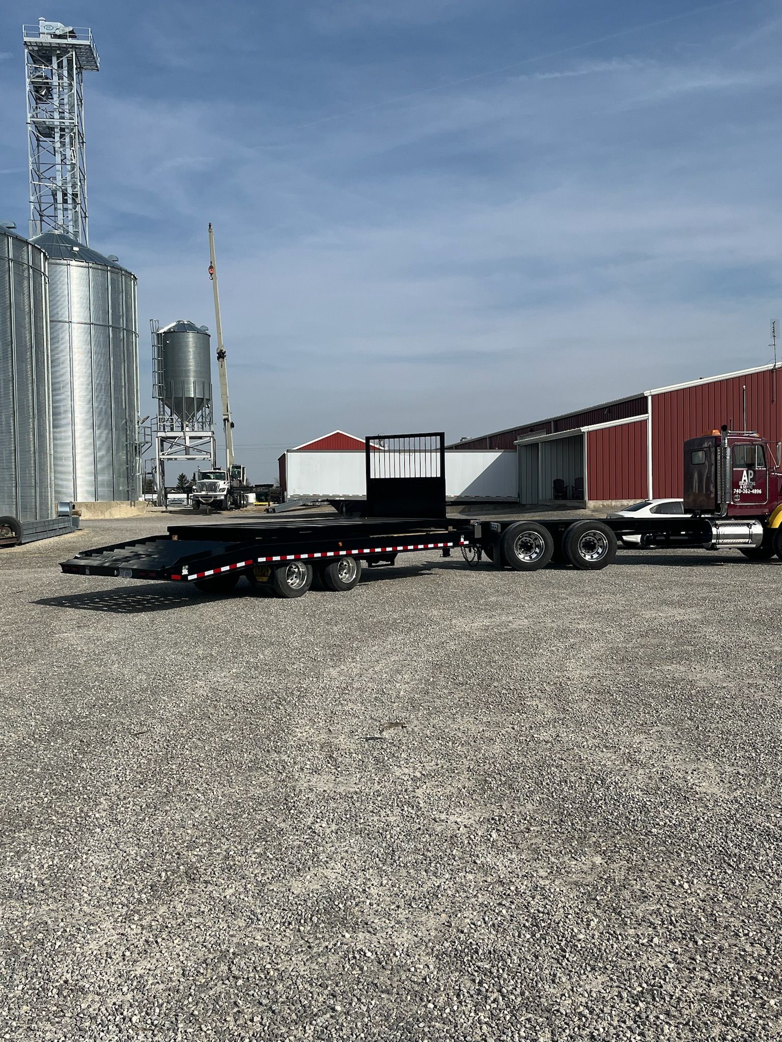 Flatbed trailer on gravel, with grain silos and a red building in the background.