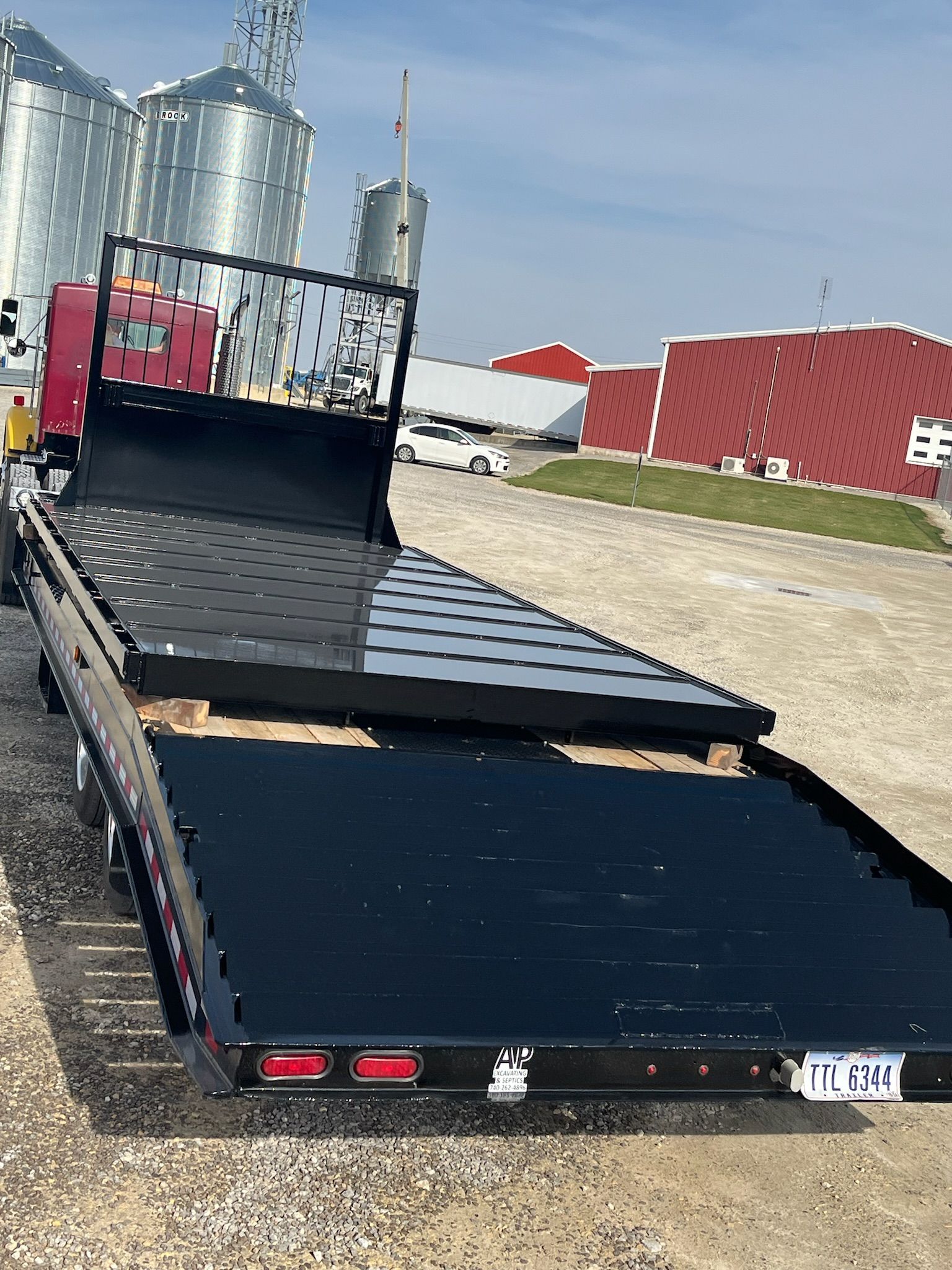 Black flatbed trailer with a red cab, parked in front of large silver silos and a brick building on a sunny day.