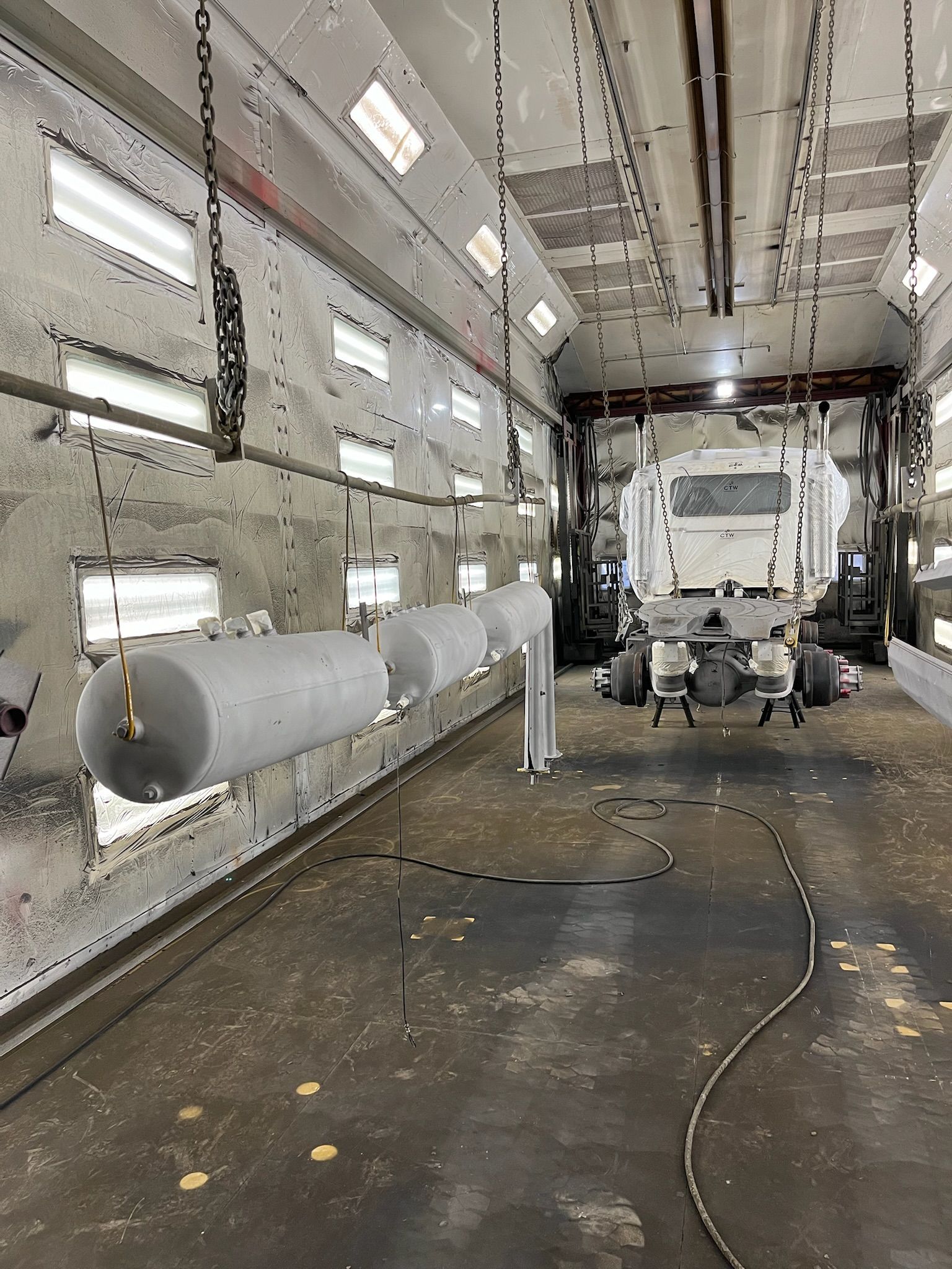Spray booth with cylindrical objects hanging, and a truck chassis visible in the background.