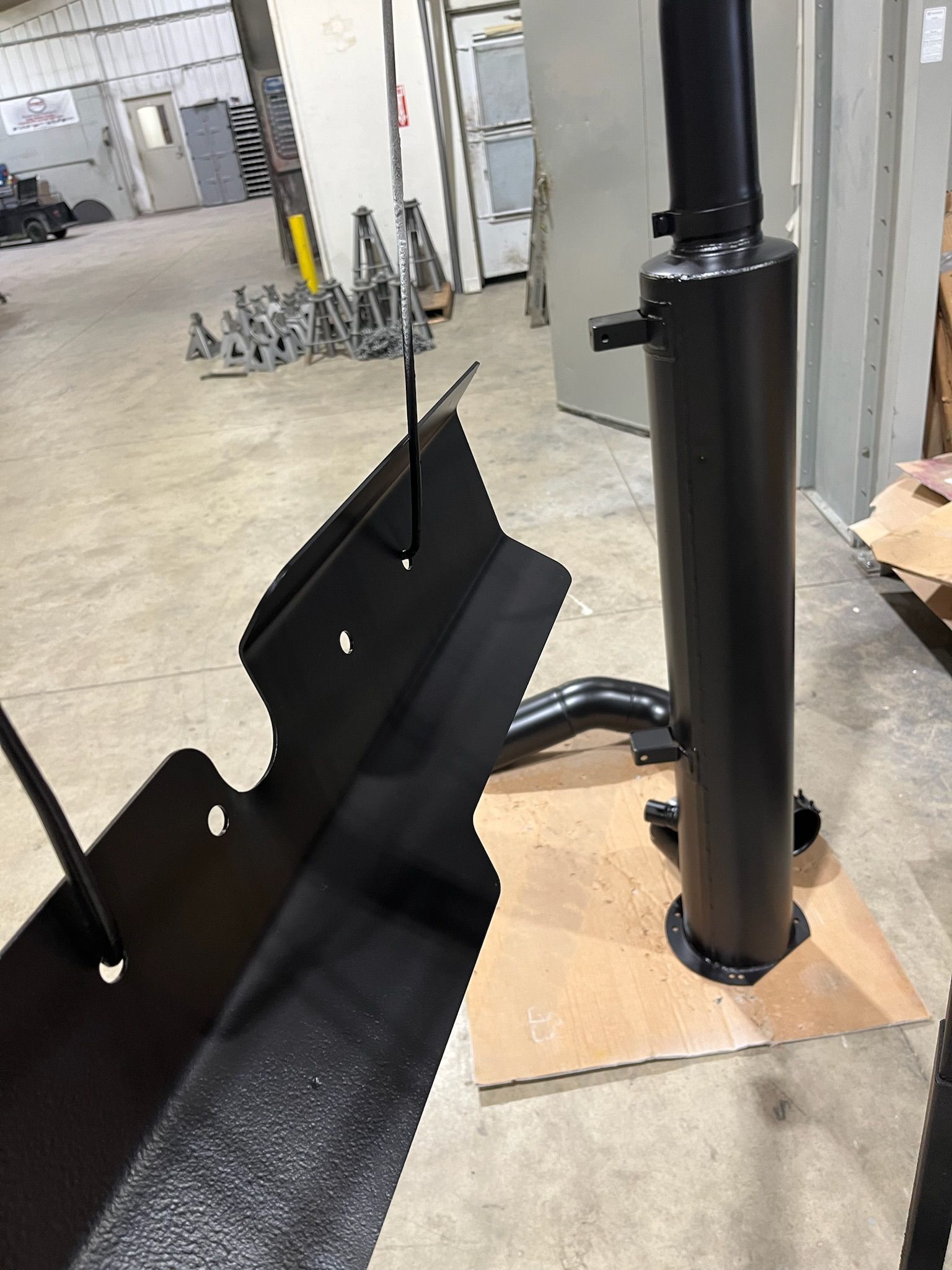 Black metal exhaust system and bracket, freshly painted, resting on cardboard in a workshop.