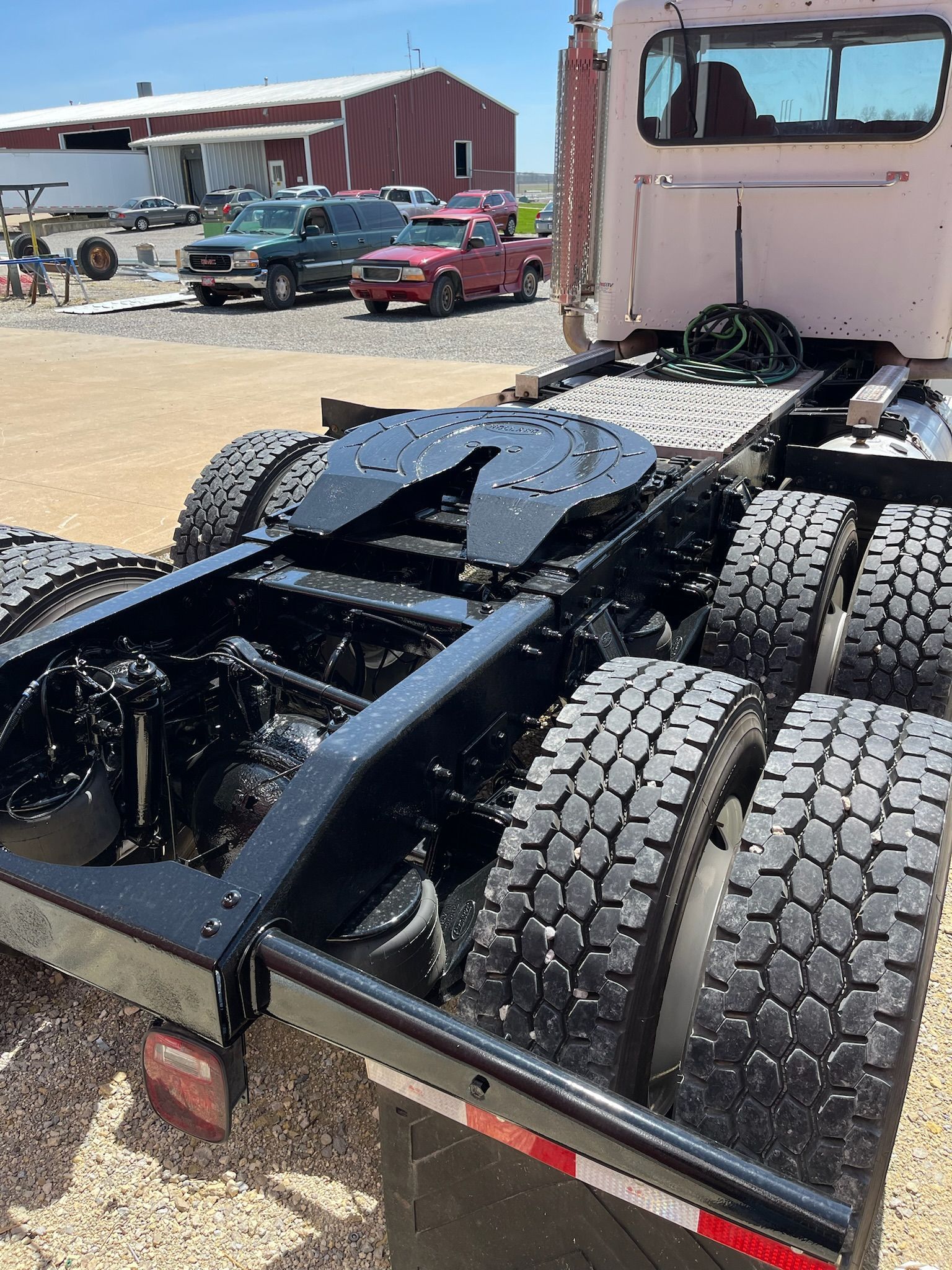 Black semi-truck chassis with dual rear wheels and fifth-wheel hitch, outdoors with vehicles and buildings.