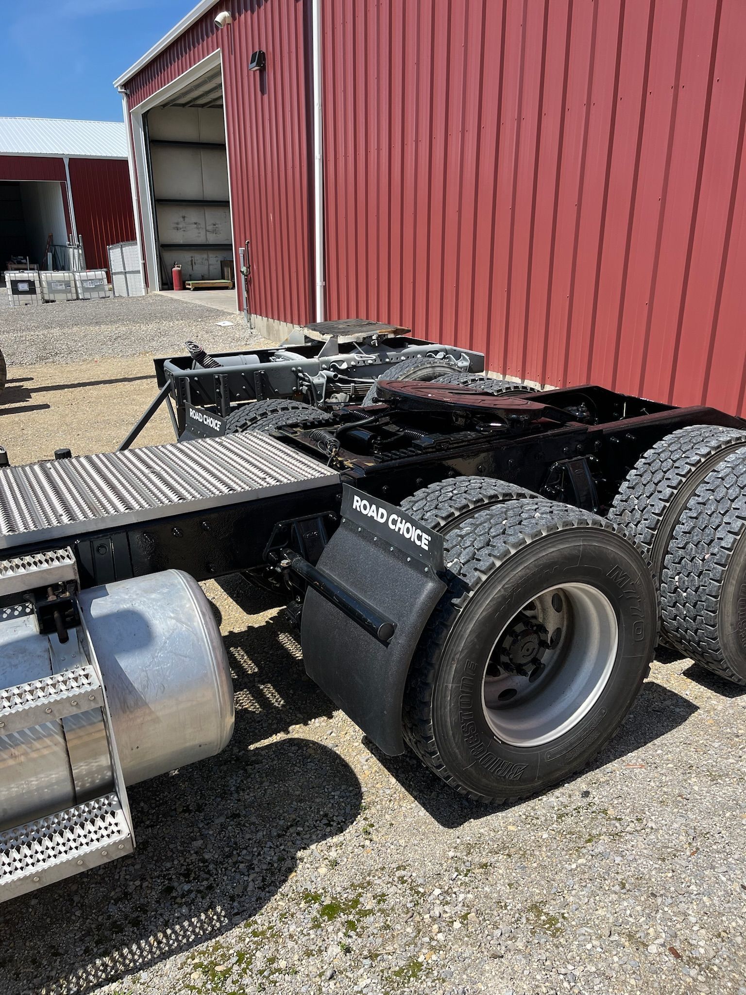 Black semi-truck chassis with tires, fuel tank, and mud flaps against a red building.
