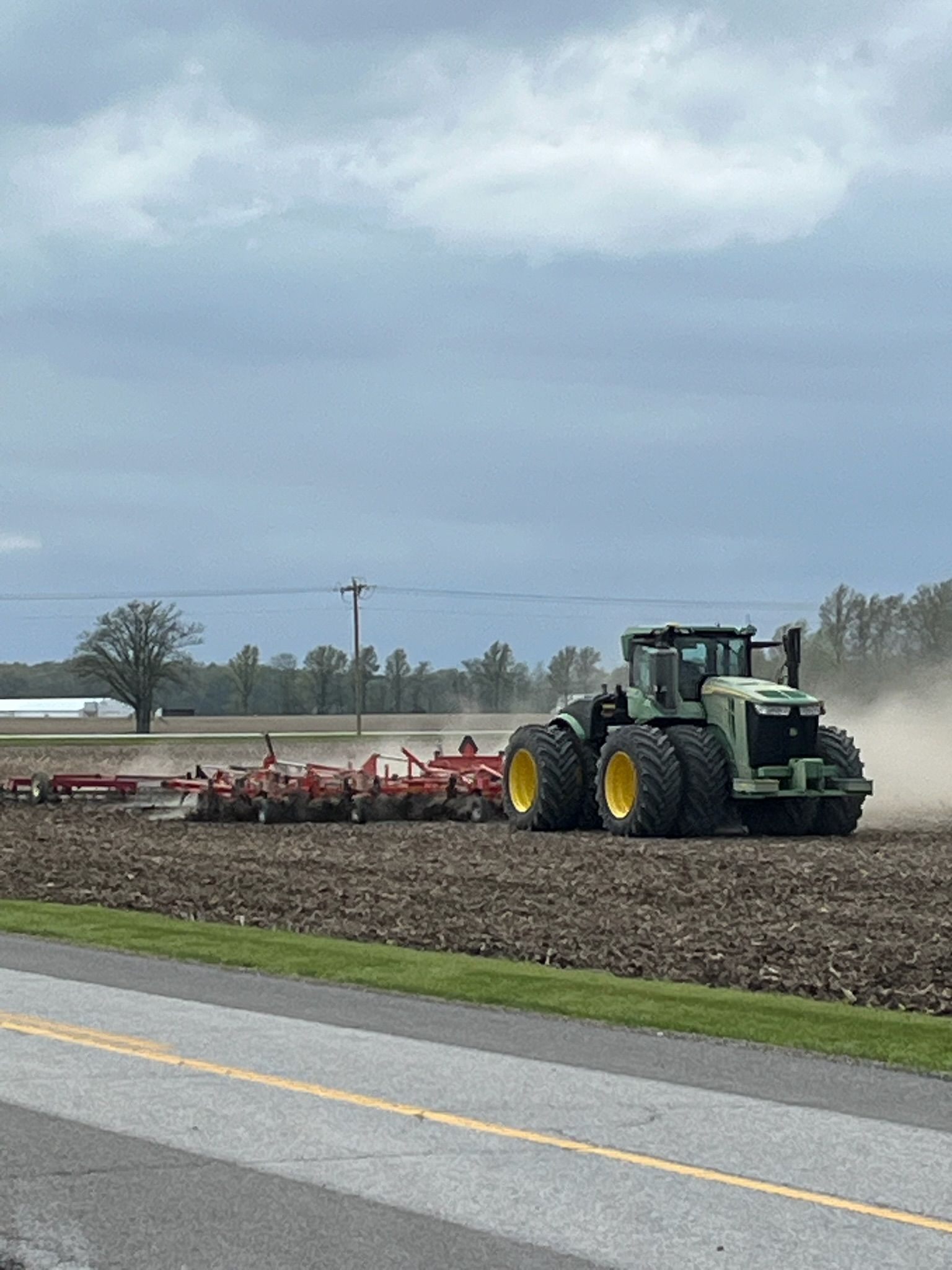 Tractor plowing a field near a road under a cloudy sky; dirt and dust in the air.