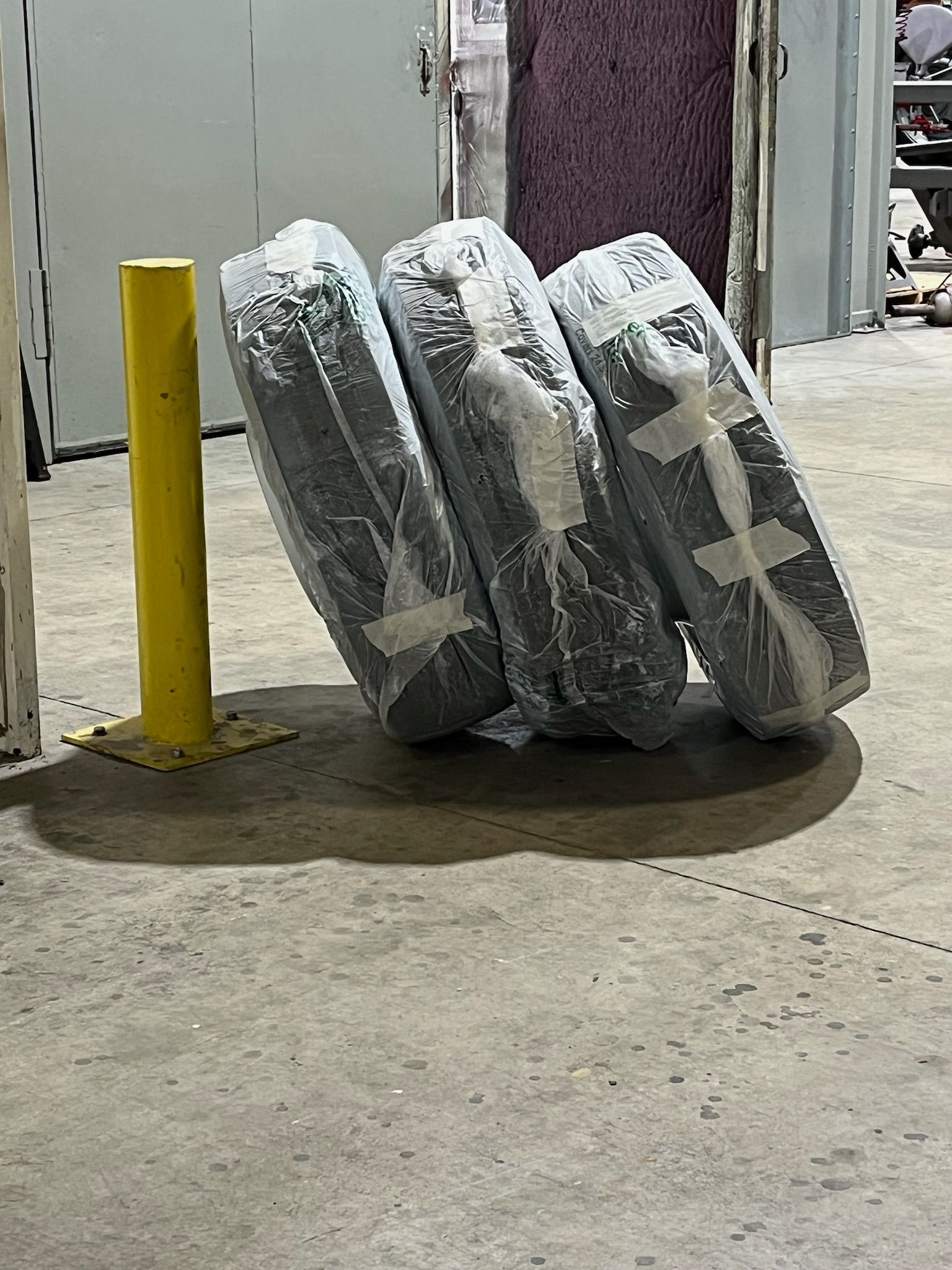 Three wrapped tires leaning against a yellow pole on a concrete floor in a warehouse.
