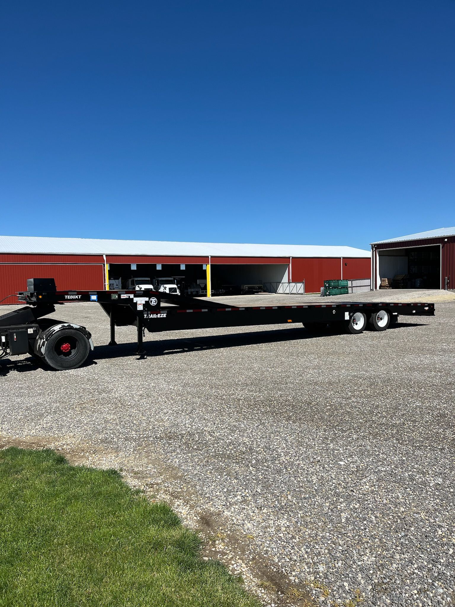 Black flatbed trailer parked on gravel, with a building in the background against a blue sky.