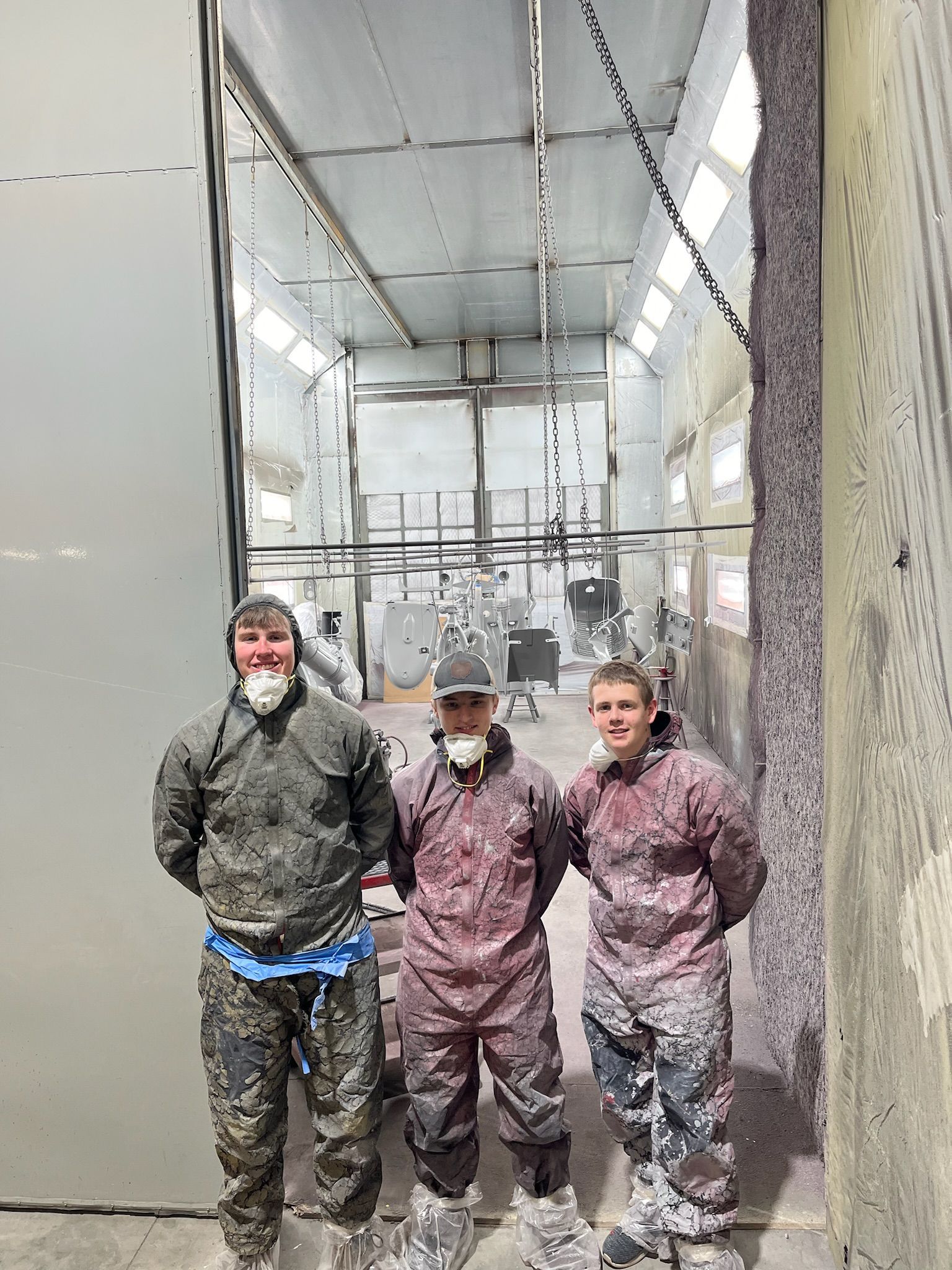 Three people in protective suits stand inside a spray booth. The booth and suits are covered in paint.